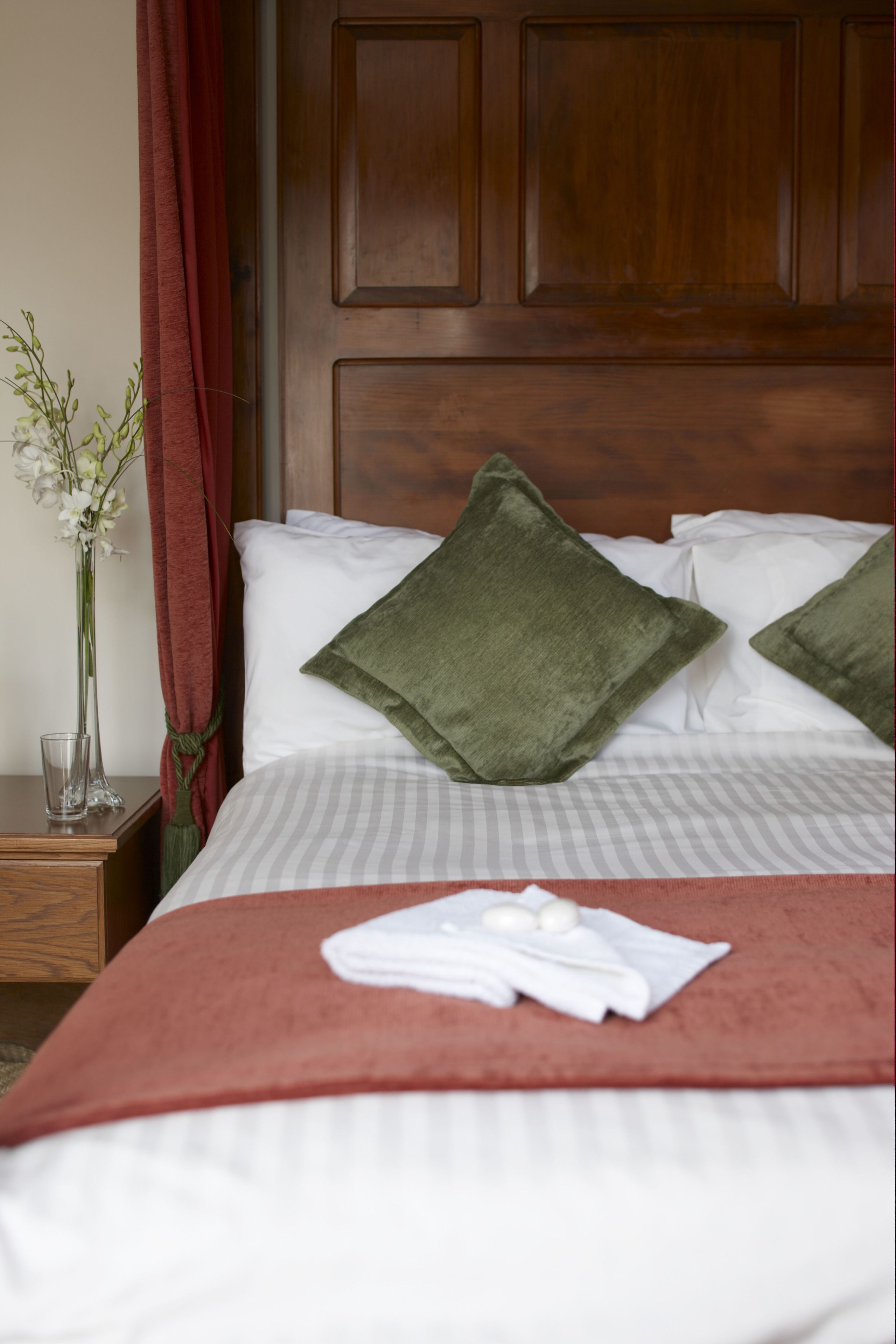 A neatly made bed with a red blanket, two green pillows, and a vase of flowers on a wooden nightstand.