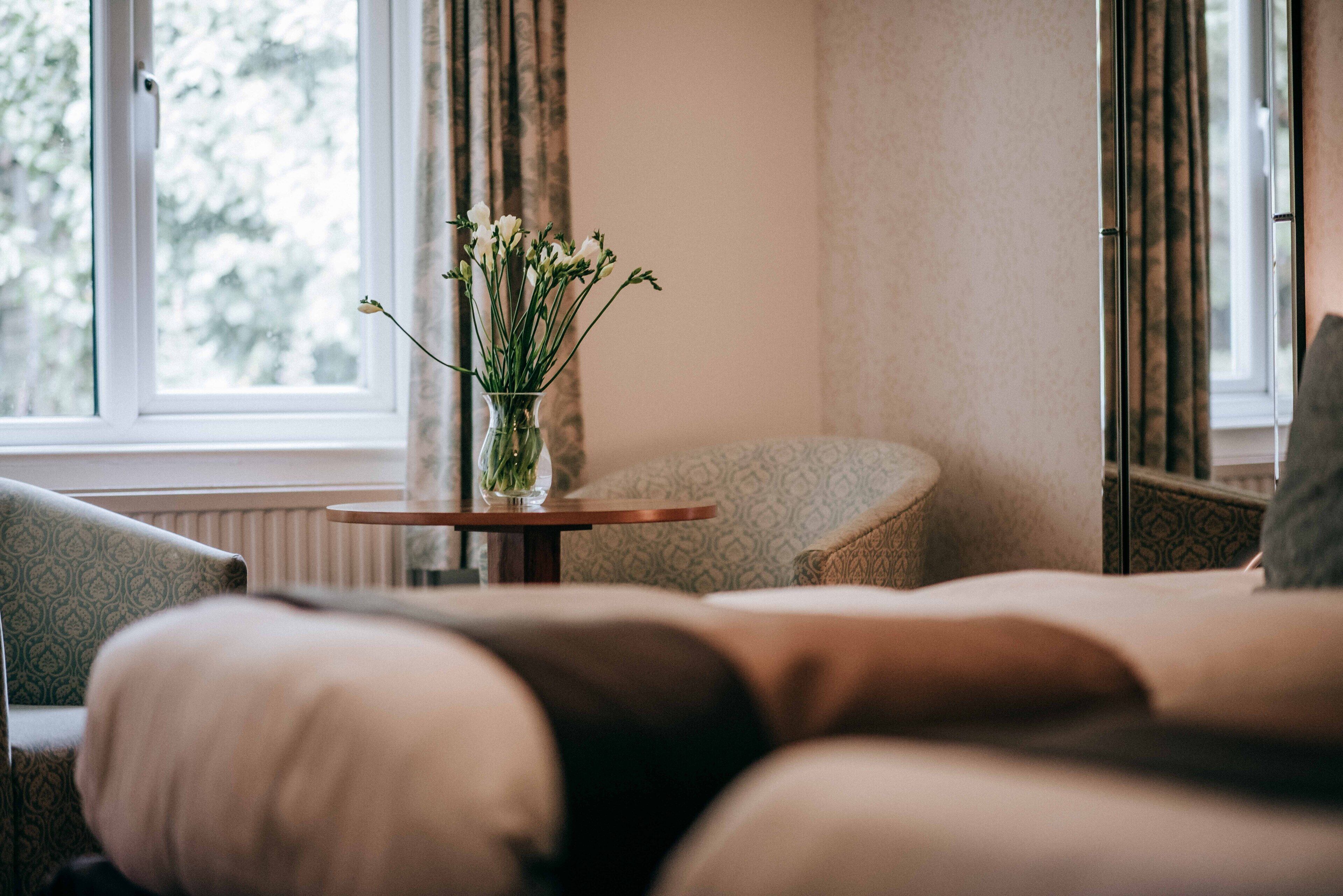 Ett mysigt rum med en soffa, ett litet runt bord med en vas med blommor och ett fönster med gardiner.