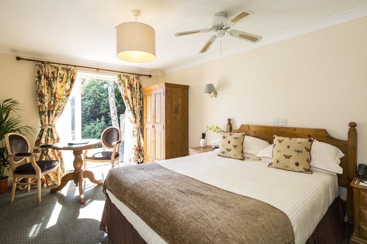 Une chambre avec un grand lit, un ventilateur de plafond, une fenêtre avec des rideaux, une petite table avec des chaises et une plante en pot.