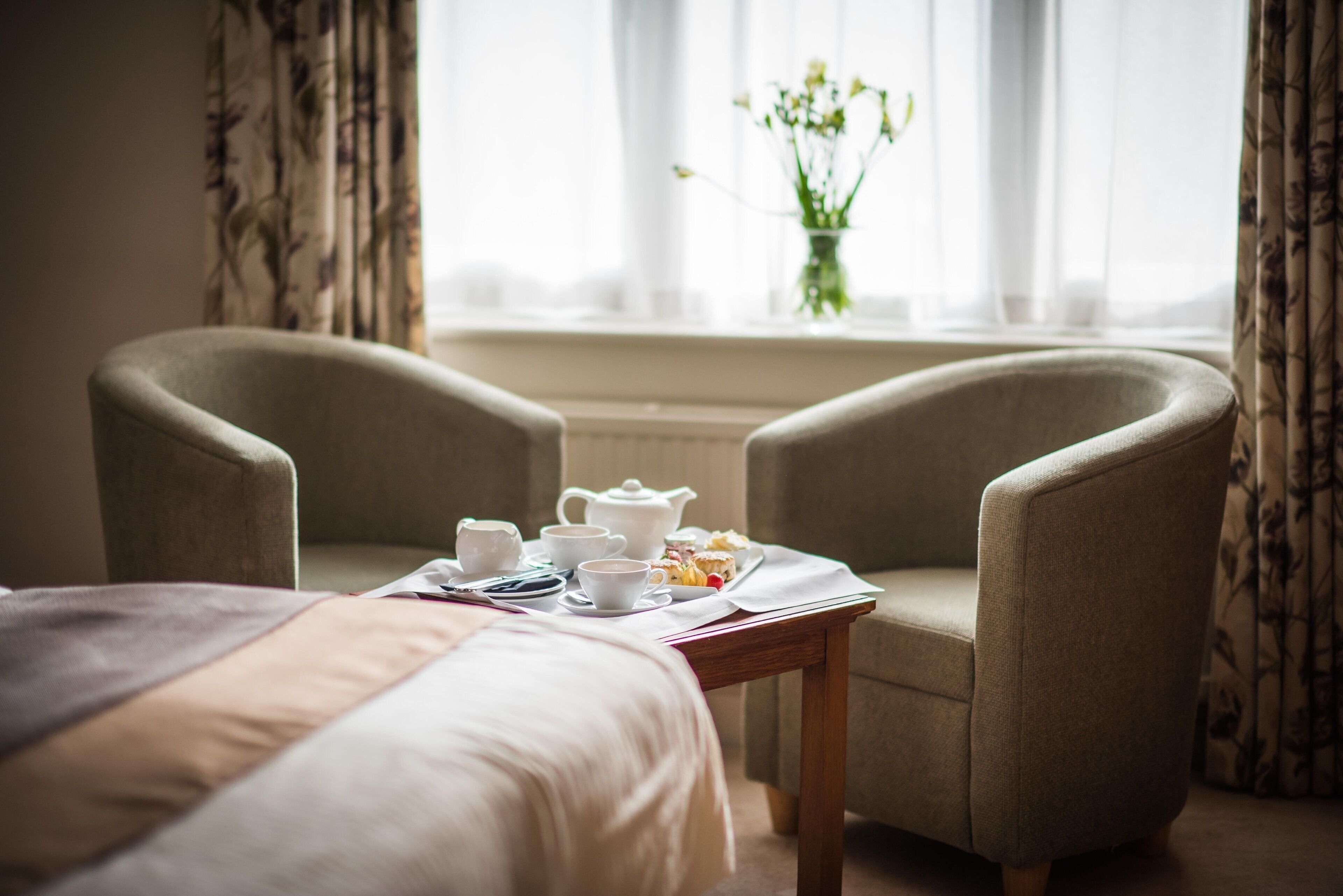 A hotel room with a bed, two armchairs, a small table with tea set and pastries, and a window with curtains and a vase of flowers.