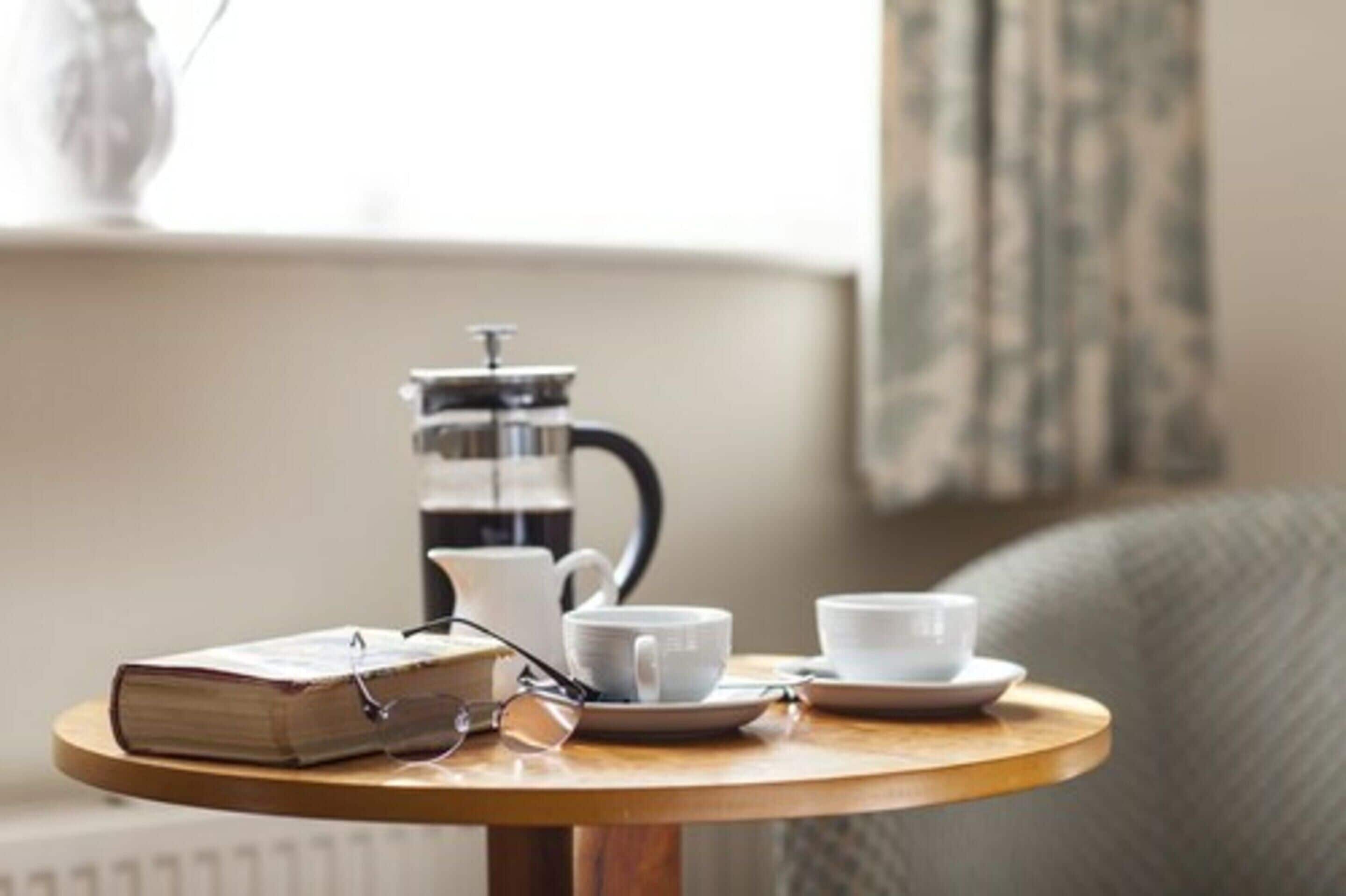 Een ronde houten tafel met een boek, glazen, een witte kop en schotel, en een koffiezetapparaat.