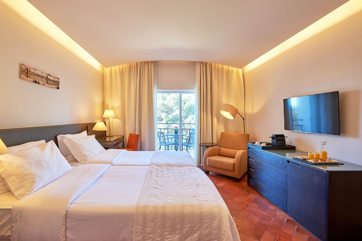 Une chambre d’hôtel avec deux lits, un bureau avec une chaise, une télévision et un balcon avec une table et des chaises.