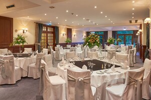 Un salón de banquetes con mesas redondas, manteles blancos y sillas adornadas con cintas de satén.