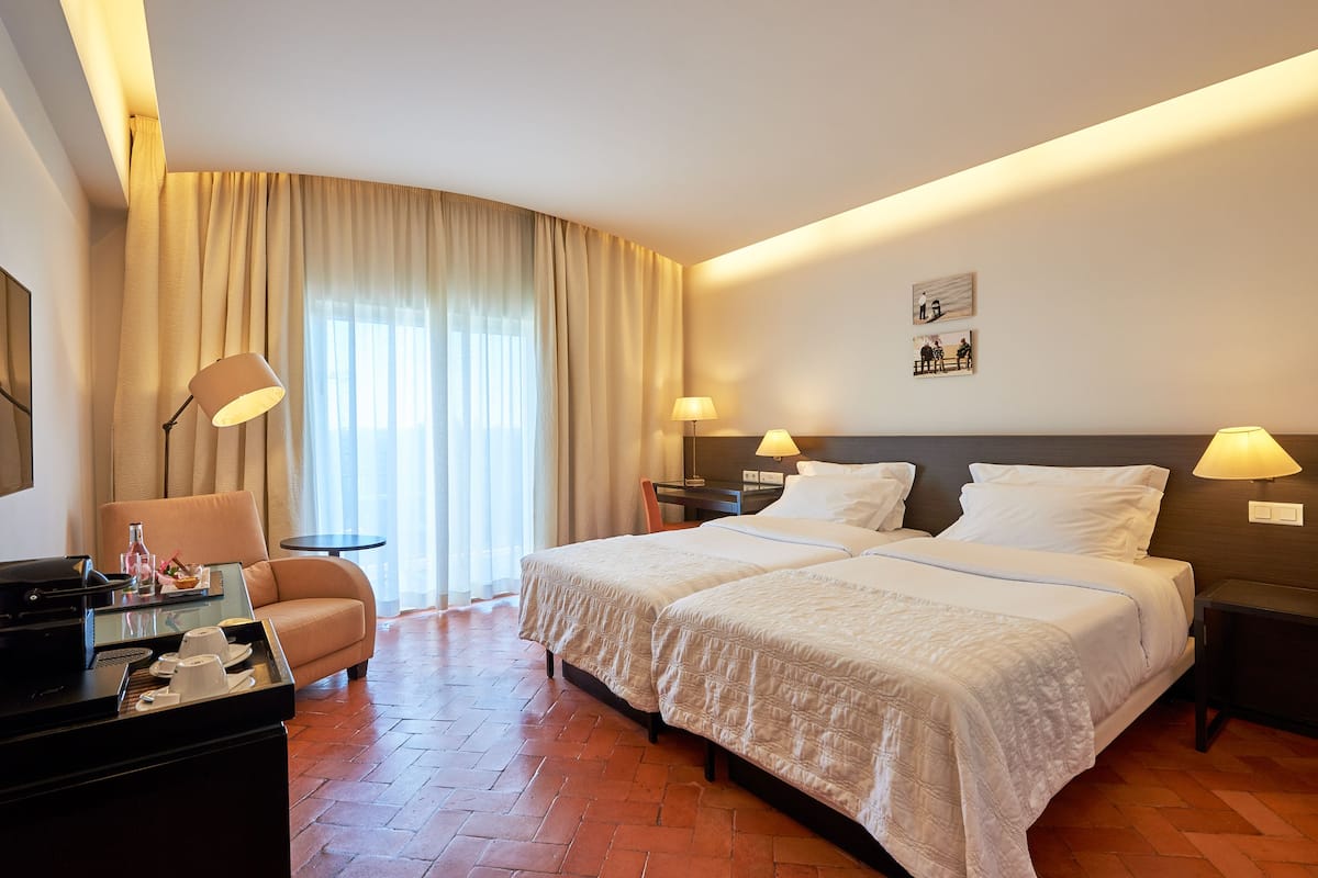 Une chambre d’hôtel avec deux lits, un canapé, un bureau avec une lampe et une grande fenêtre avec des rideaux.