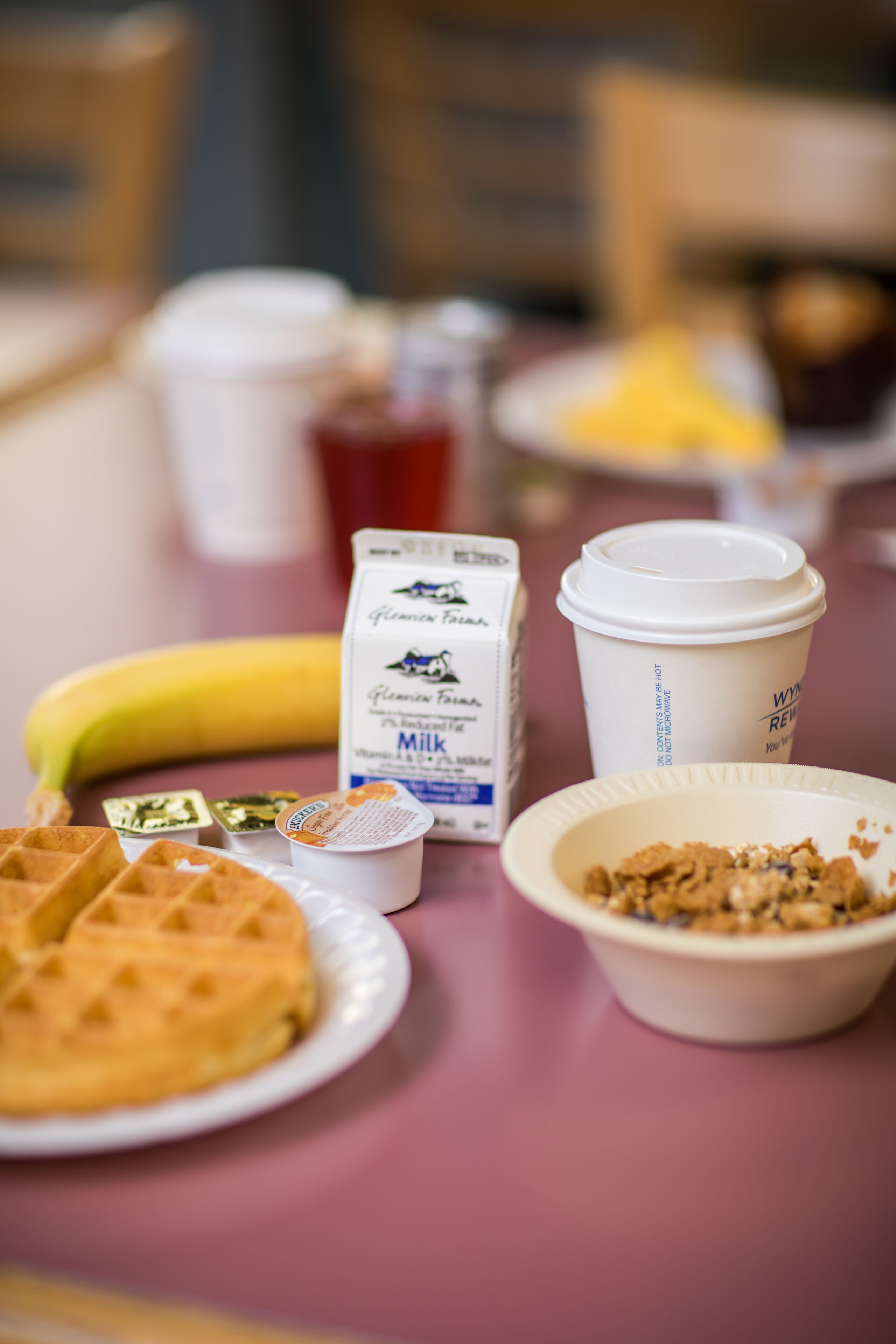 A table with a banana, a carton of milk, a coffee cup, a waffle, and a bowl of cereal.