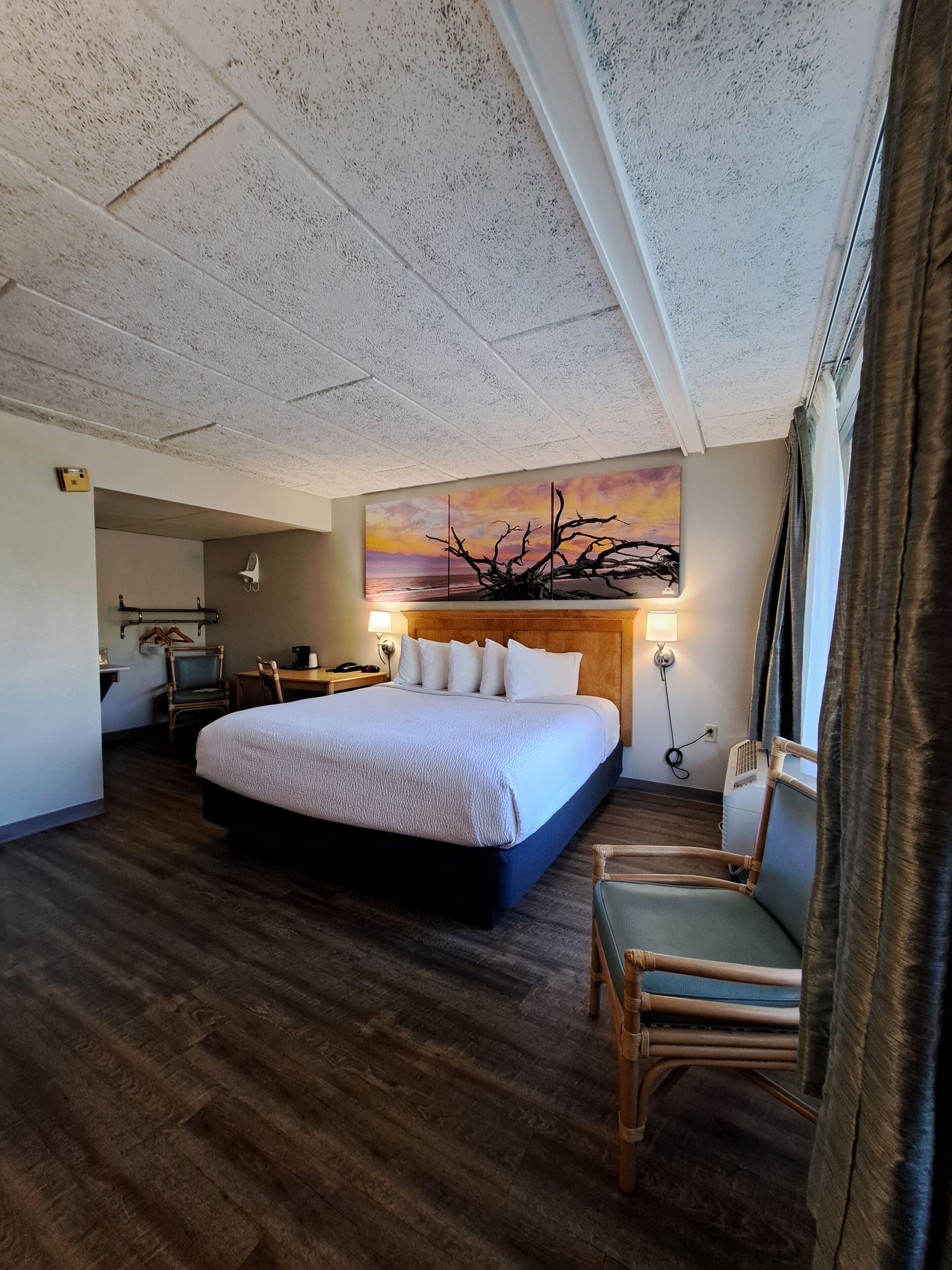 A hotel room with a large bed, a desk with a chair, a painting on the wall, and a window with curtains.