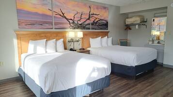 Habitación de hotel con dos camas, cabecera de madera, lámparas de noche, mesitas de noche, una pequeña mesa, un espejo y un cuadro de un atardecer sobre el océano.