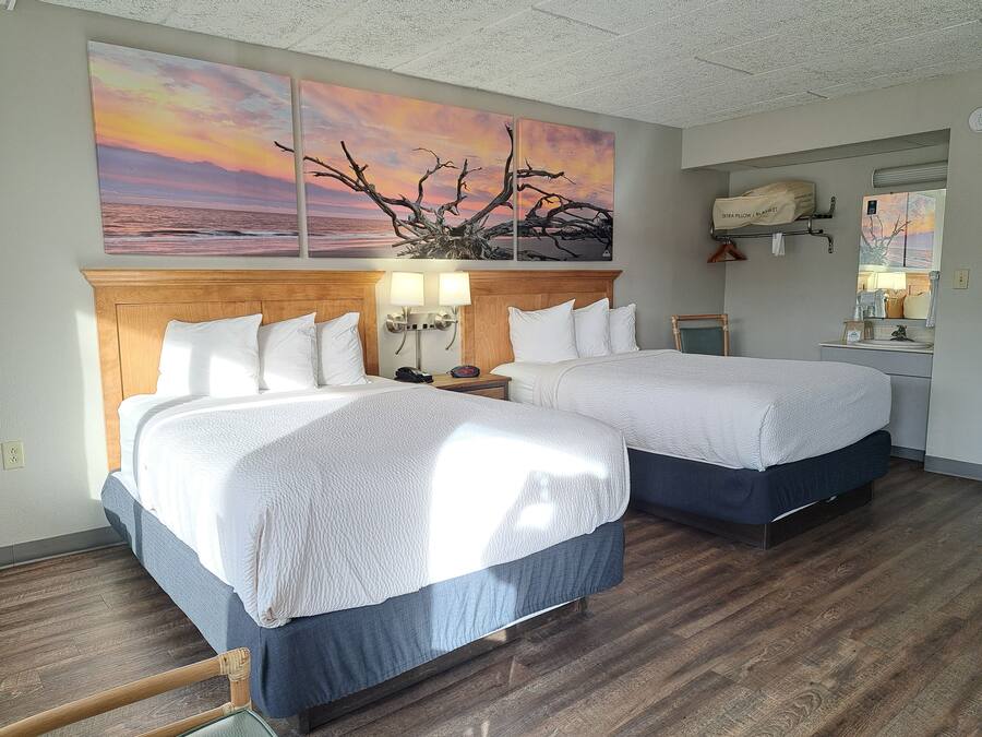 Habitación de hotel con dos camas, cabecera de madera, lámparas de noche, mesitas de noche, una pequeña mesa, un espejo y un cuadro de un atardecer sobre el océano.