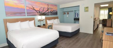 A hotel room with two beds, a wooden floor, a painting of a sunset, and a kitchenette area.