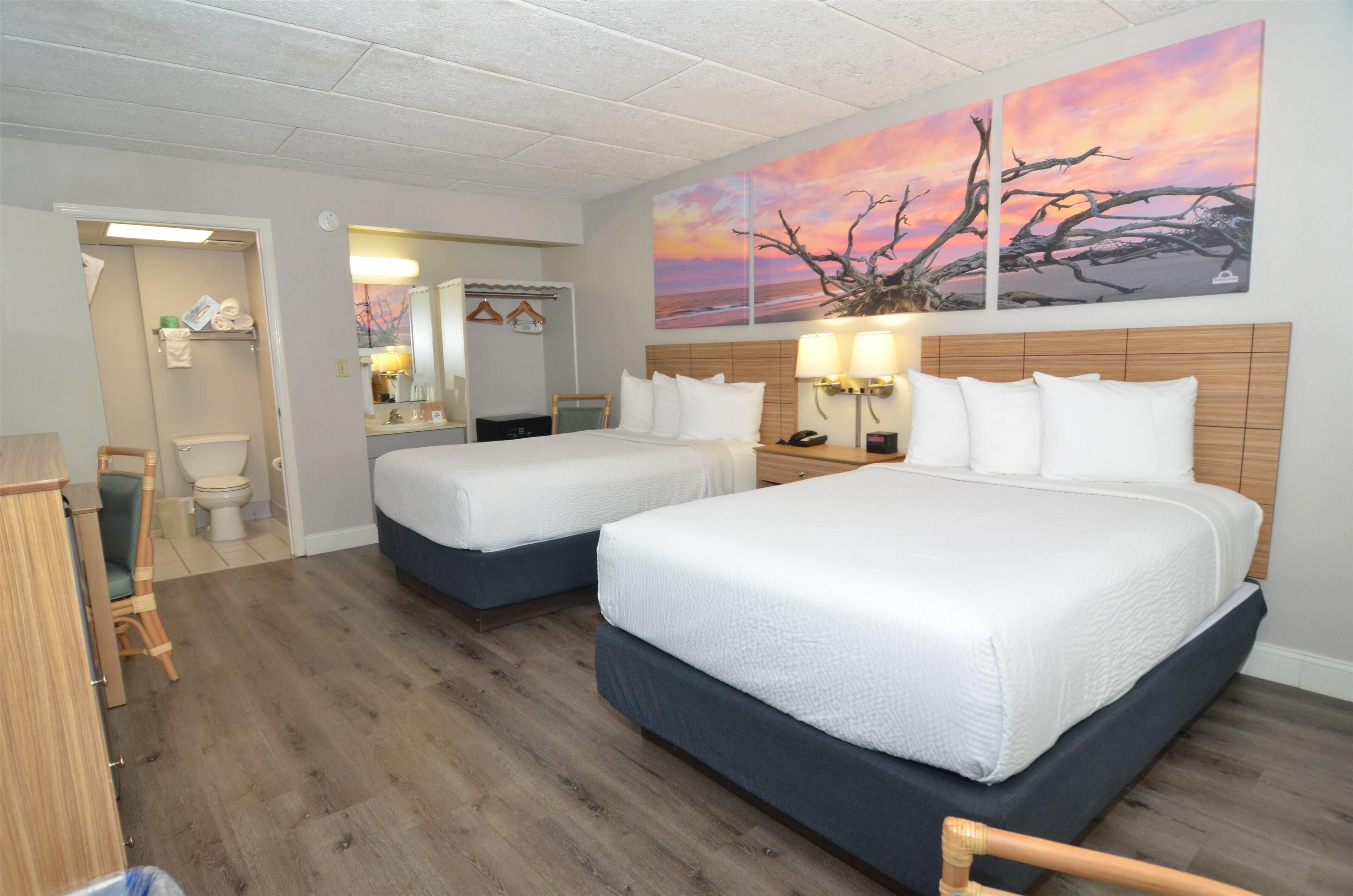 A hotel room with two beds, a wooden headboard, a bathroom with a toilet and sink, and a sunset painting on the wall.