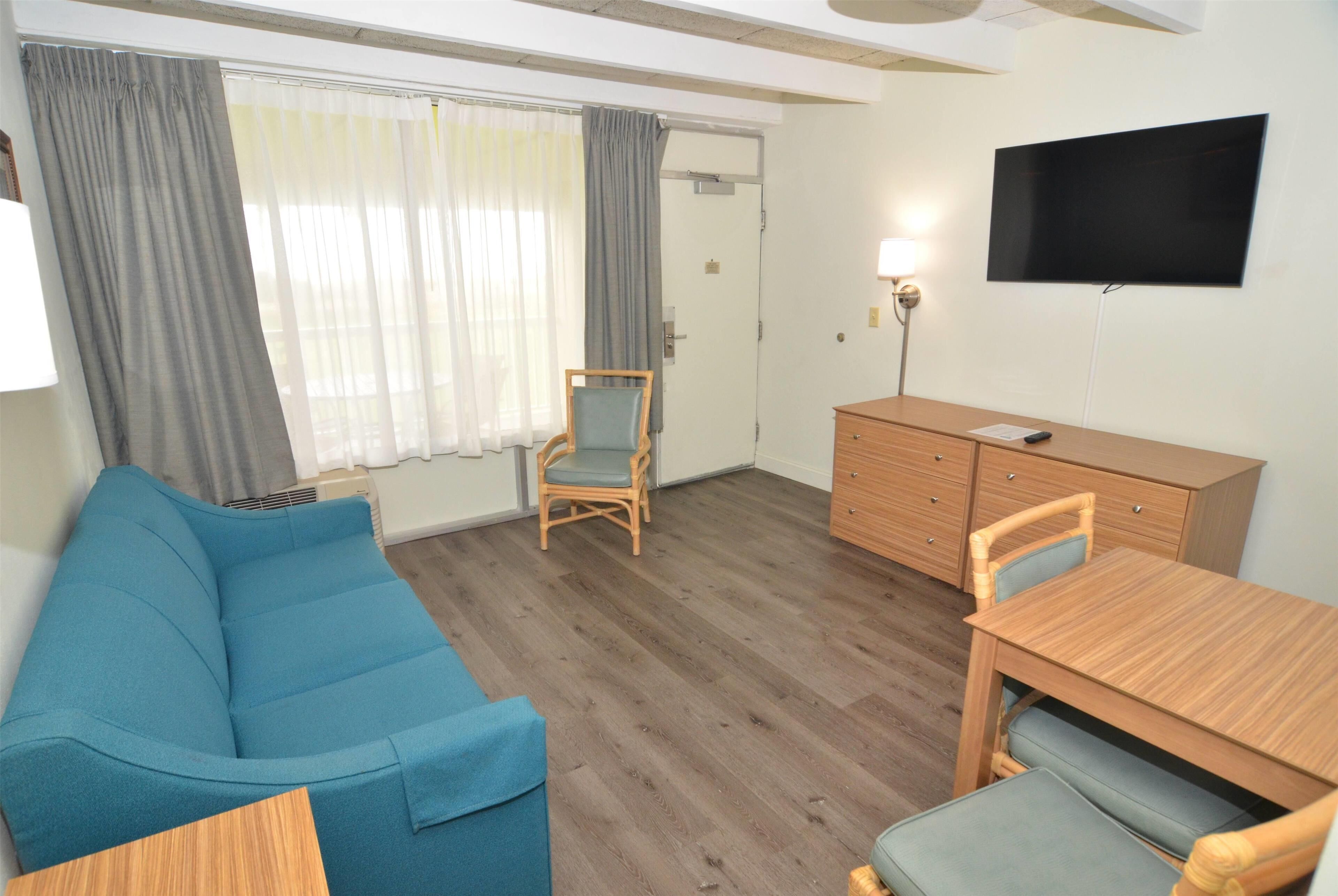 A room with a blue sofa, a wooden dining table with chairs, a TV on the wall, and a wooden cabinet.