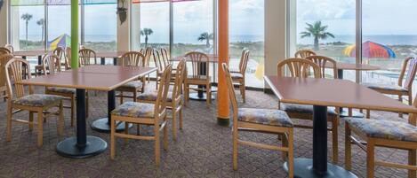 A dining area with tables and chairs, large windows with a view of the beach, and a colorful wall decoration.
