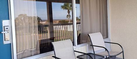A covered outdoor seating area with two chairs and a table, a sliding glass door, and a view of the beach.