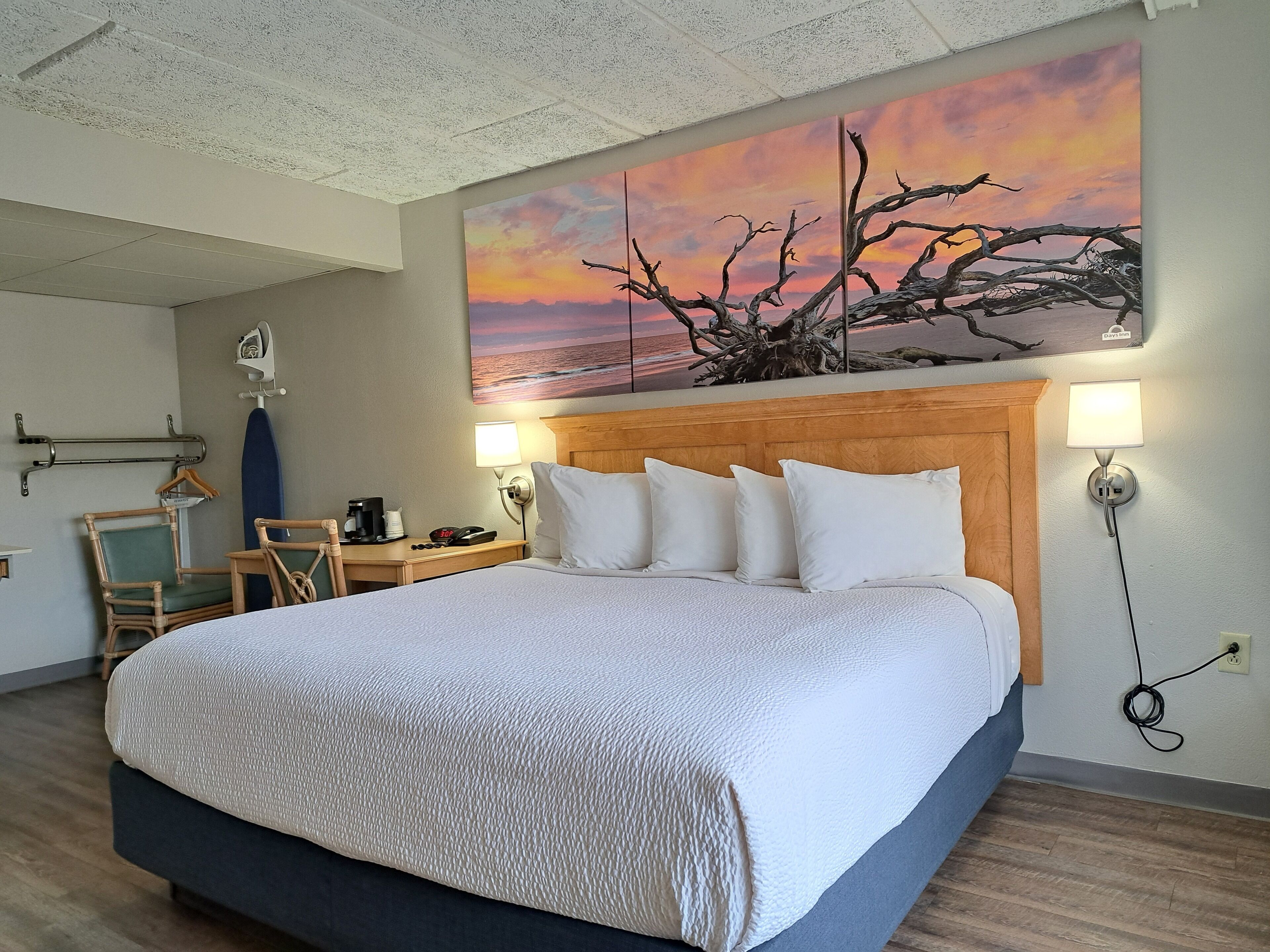 Een hotelkamer met een groot bed, een houten hoofdbord, een nachtkastje met een lamp, een stoel en een schilderij van een zonsondergang.