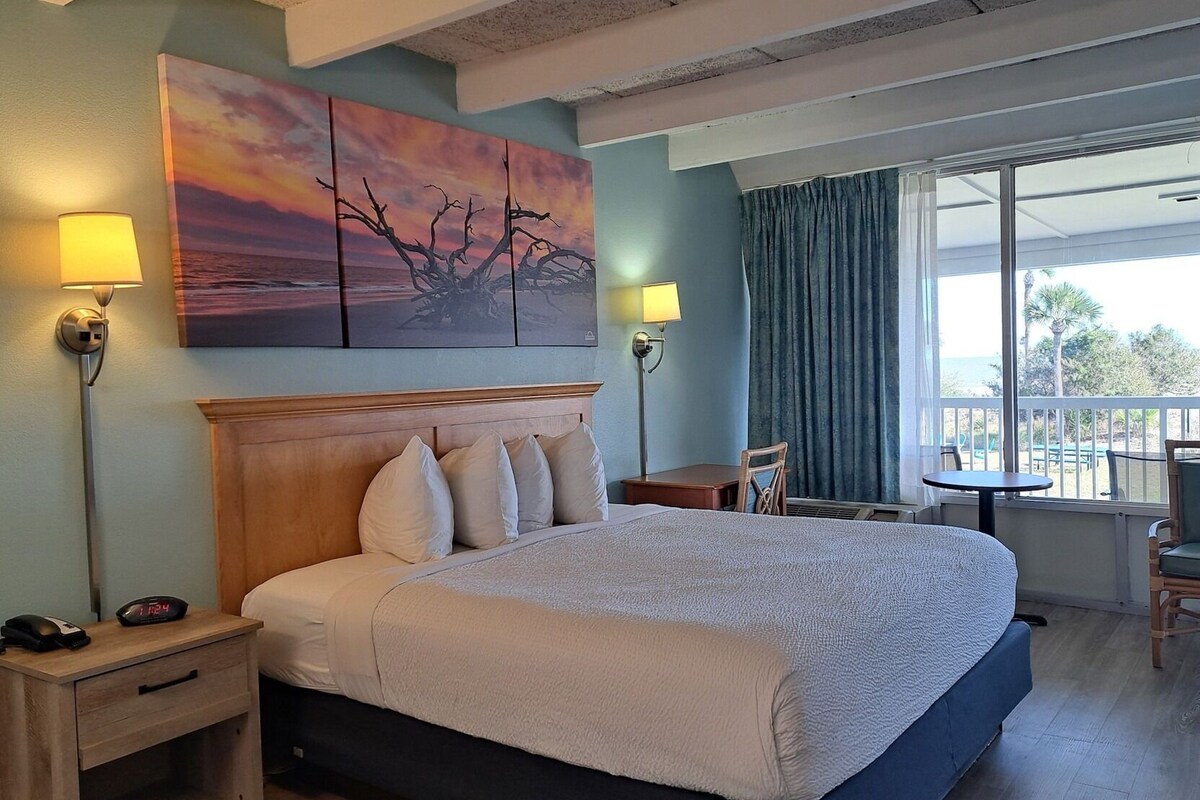 A hotel room with a large bed, bedside lamps, a painting, a view of the ocean, and a balcony with chairs.