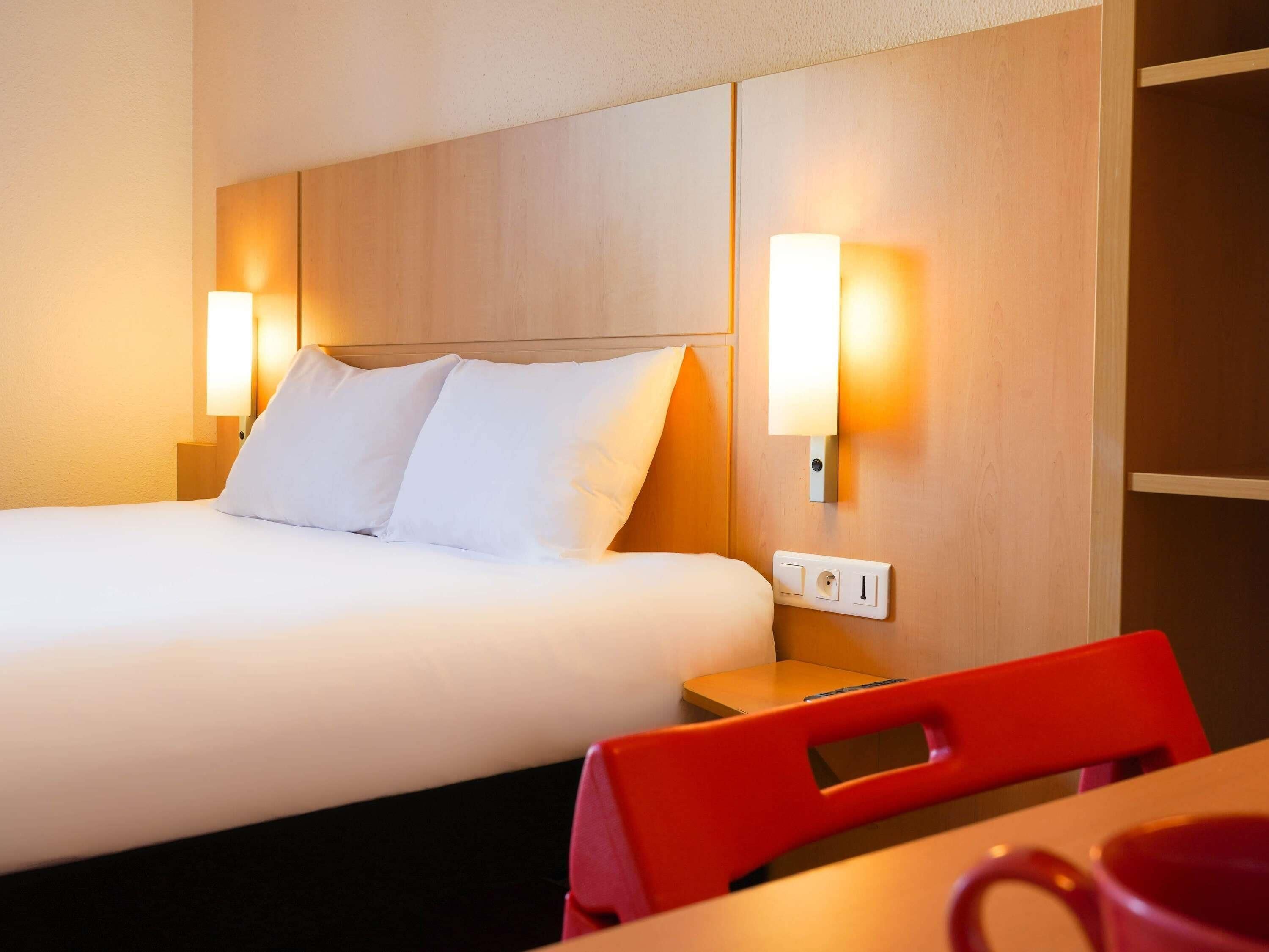 A hotel room with a bed, bedside lamps, a nightstand, a red chair, and a table with a red mug.