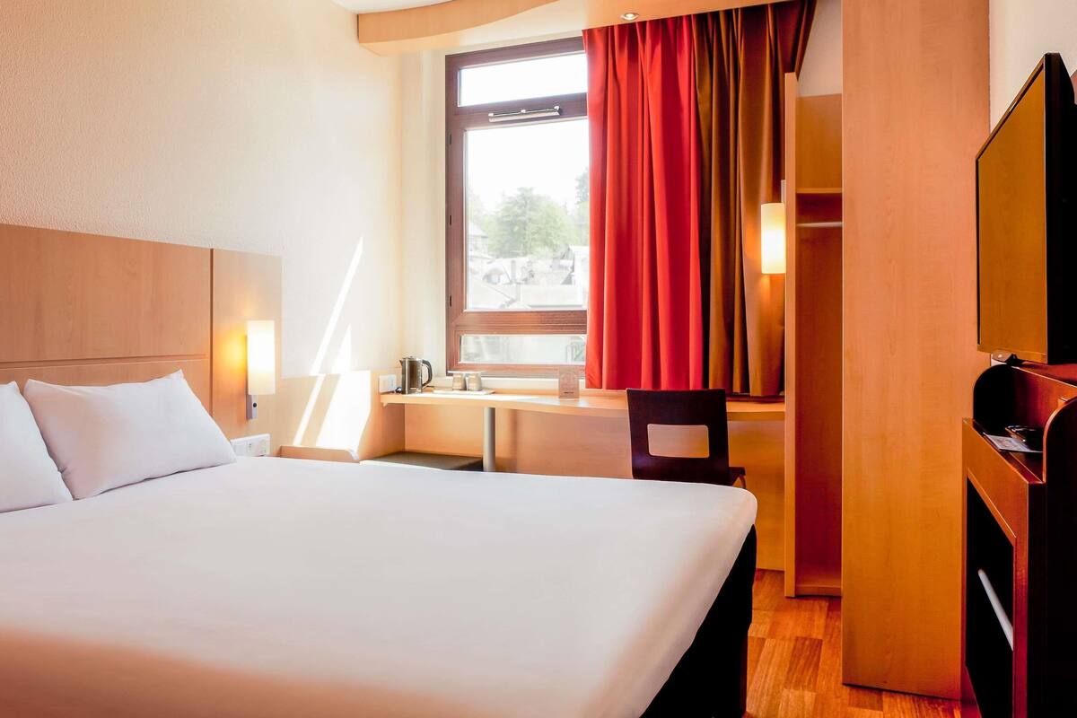 A hotel room with a bed, a desk with a lamp, a window with curtains, and a wardrobe.