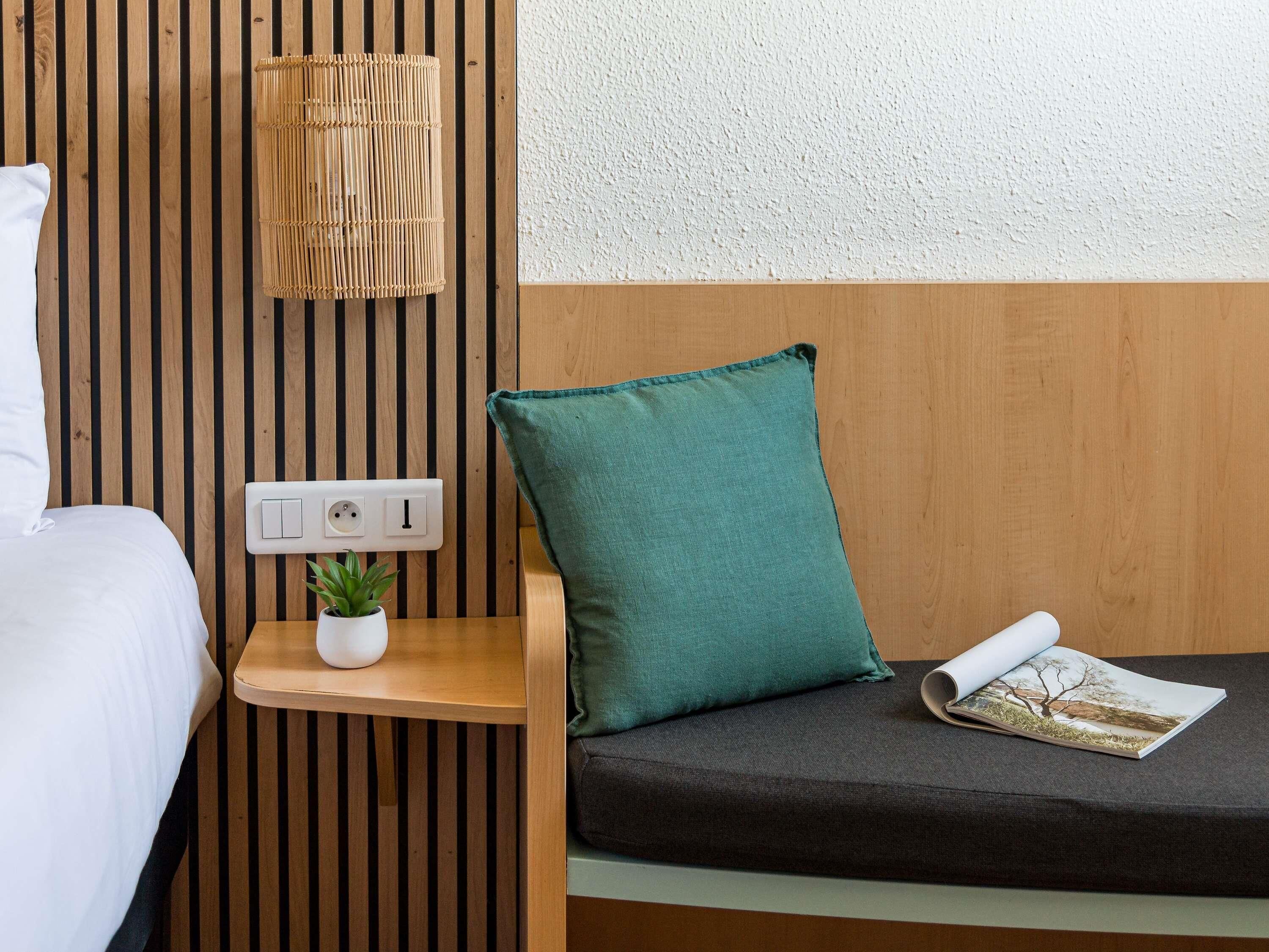 Una habitación con cama, una mesita de noche con toma de corriente, una planta pequeña en maceta, un almohadón verde y una revista.