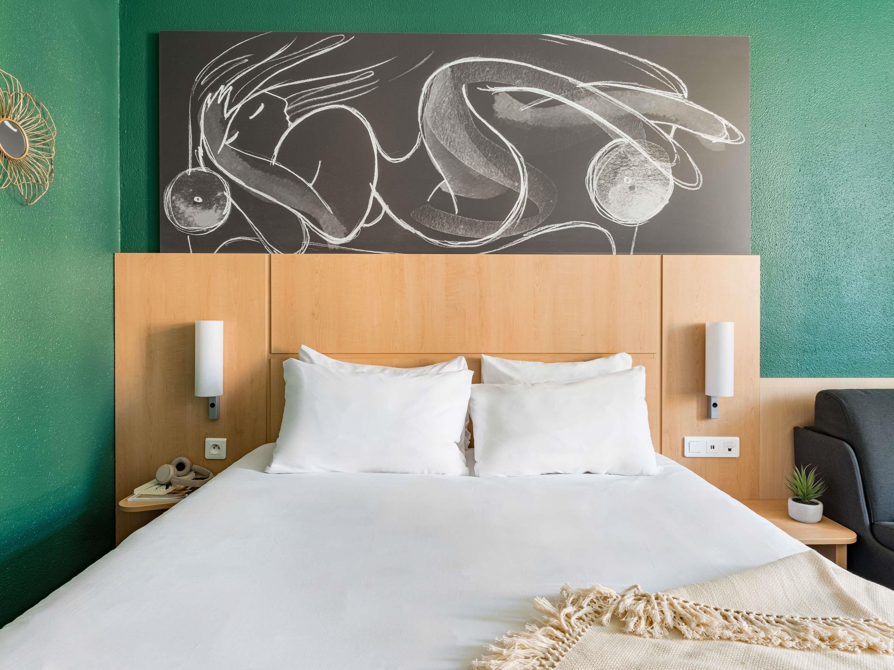 A hotel room with a bed, white pillows, a wooden headboard, two wall-mounted lights, a small bedside table with a phone and a plant, and a wall art piece above the bed.