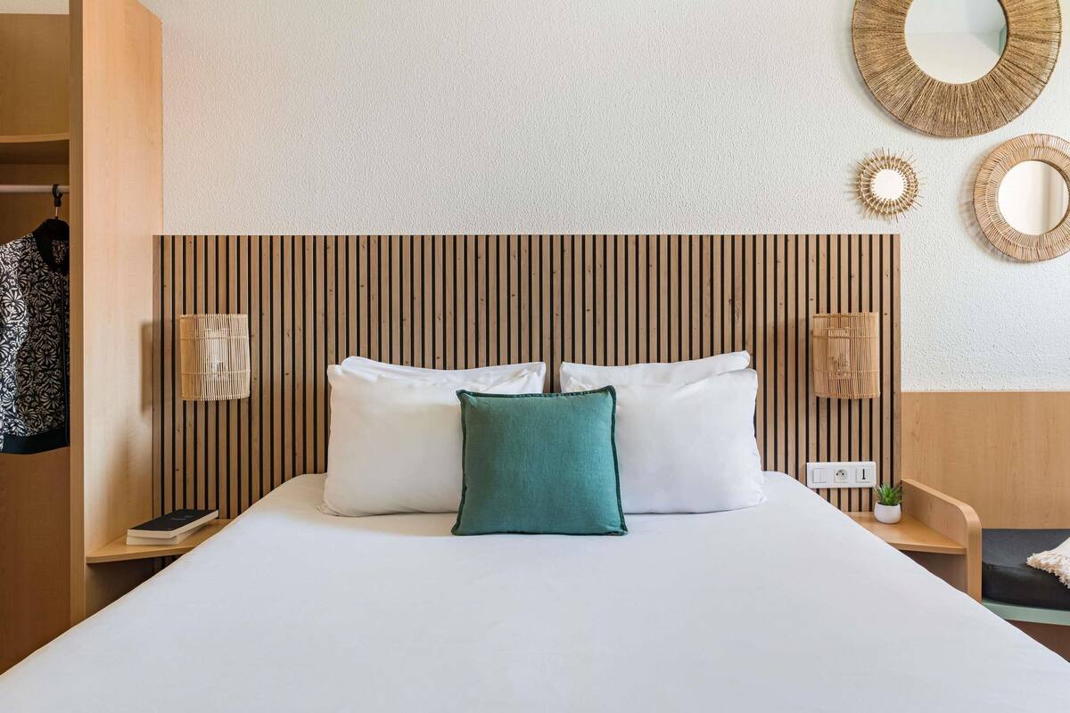 A hotel room with a bed, a bedside table with a lamp, a wall-mounted mirror, and a wooden headboard.