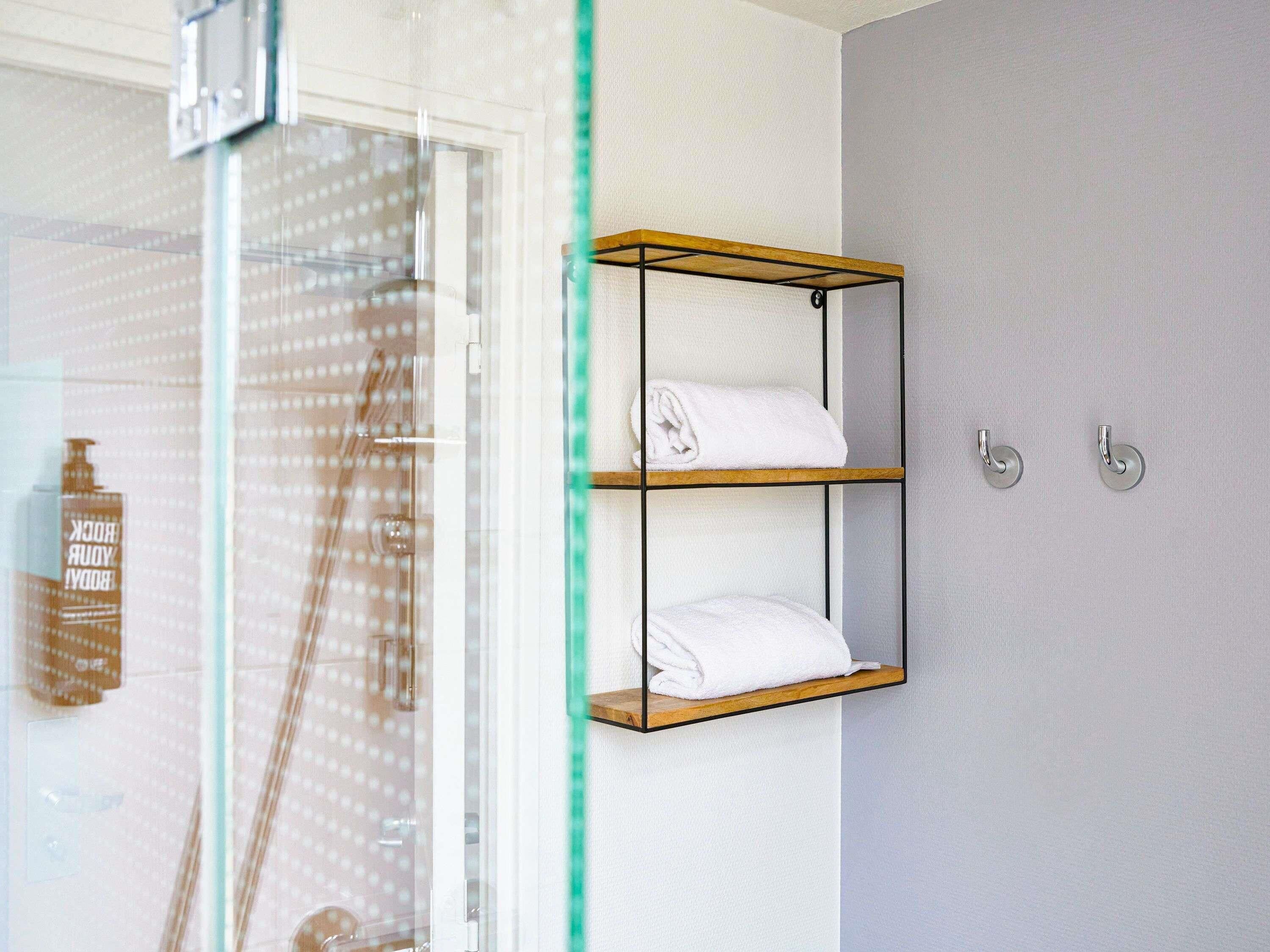 Une salle de bain avec une porte de douche en verre, une étagère en bois avec des serviettes et une bouteille de shampoing.