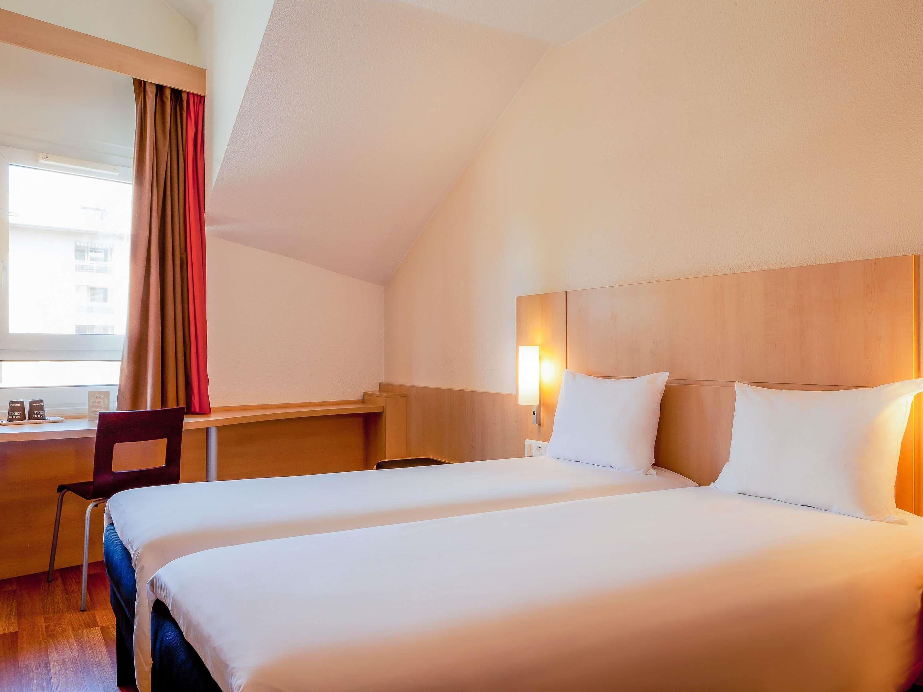 A hotel room with two beds, a desk, a chair, a nightstand, and a window with curtains.