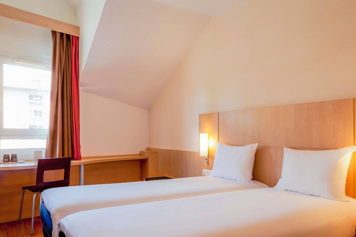 A hotel room with two beds, a desk, a chair, a nightstand, and a window with curtains.