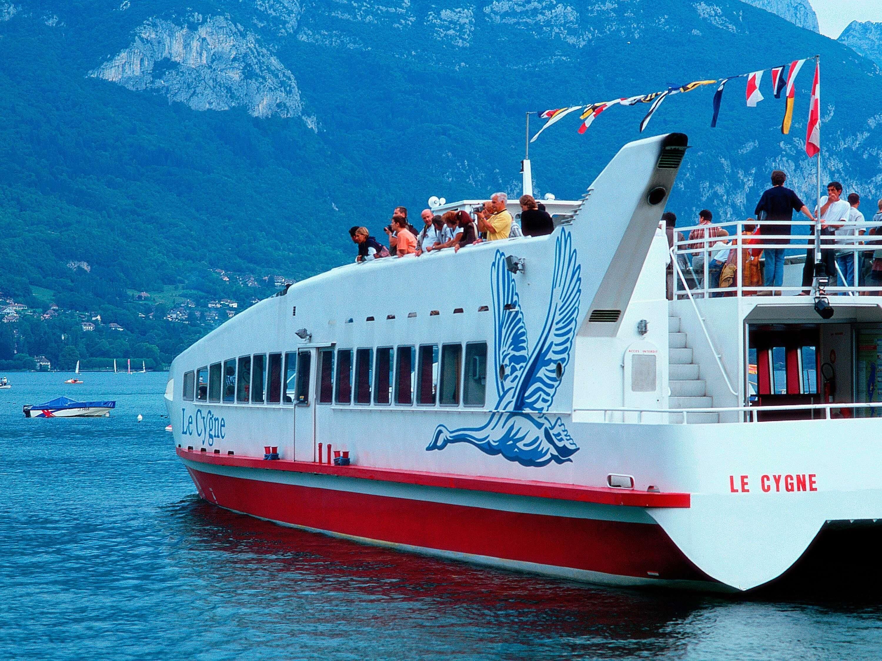 A boat with 'Le Cygne' written on the side, passengers on deck, and a mountainous backdrop.