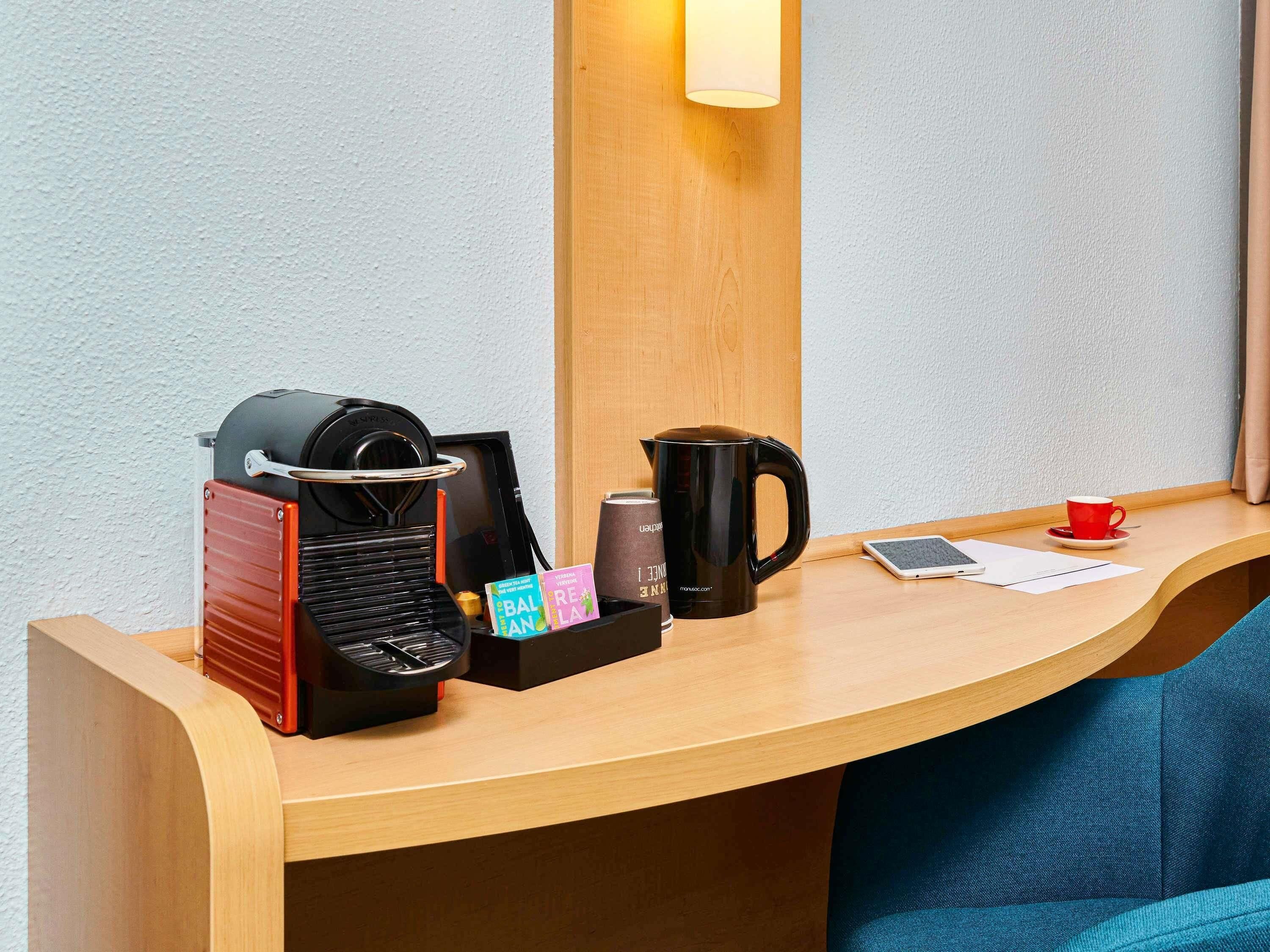 A hotel room desk with a coffee maker, a mug, a smartphone, and a tablet.