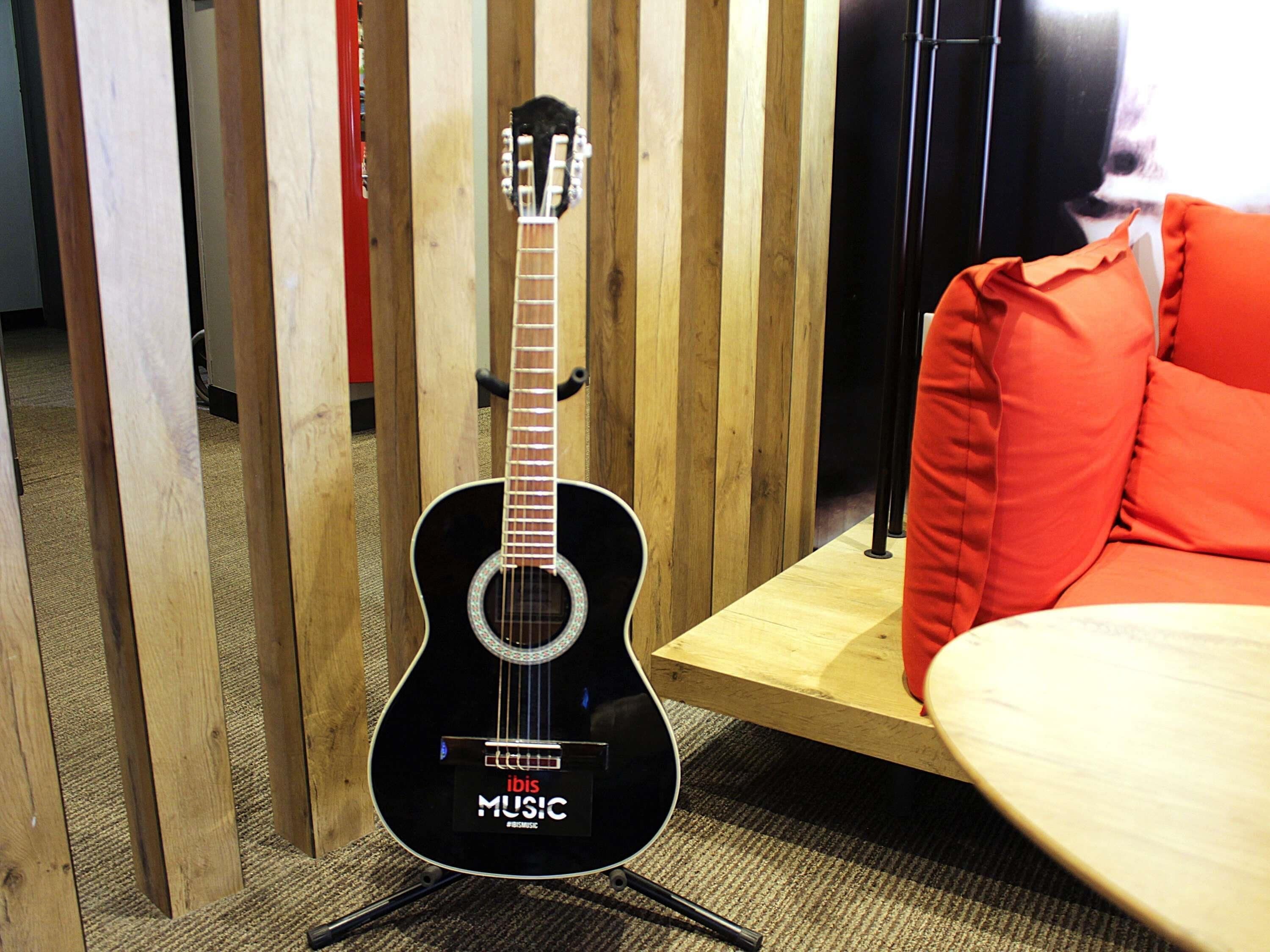 A black acoustic guitar with a 'MUSIC' label on a stand in a room with wooden slats, a red cushion, and a wooden table.