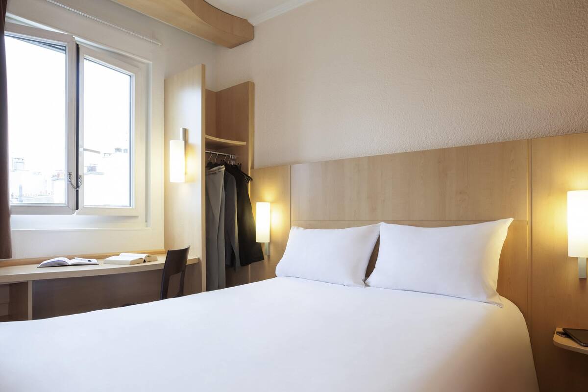 A hotel room with a bed, bedside table, lamp, and a window with curtains.