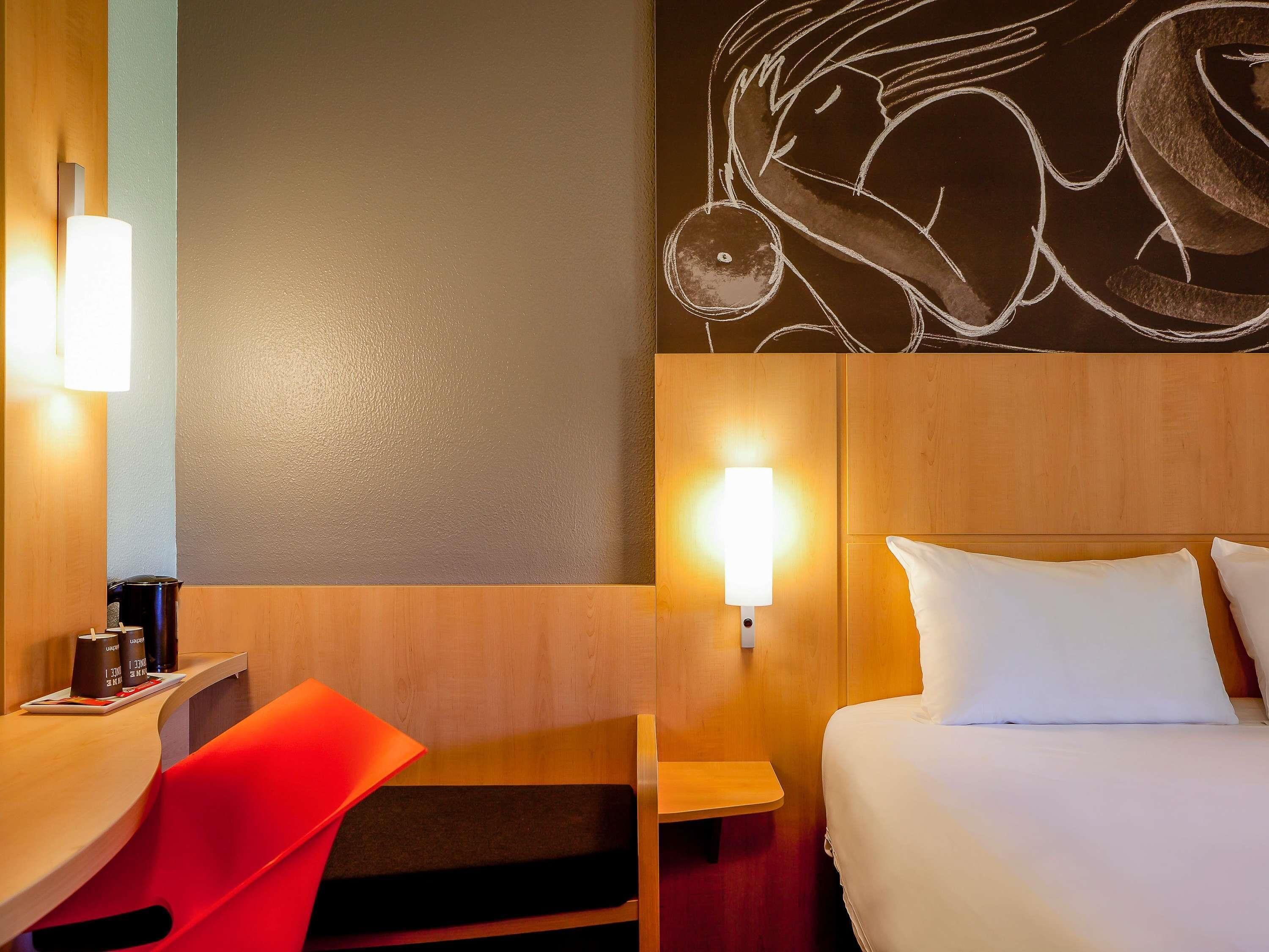 A hotel room with a bed, a desk with a chair, a wall-mounted lamp, and a wall art piece.