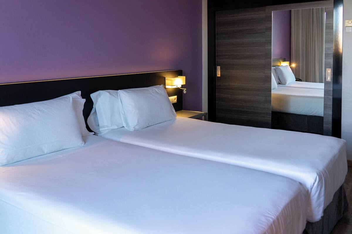A hotel room with a large bed, a nightstand with a lamp, and a mirror reflecting the bed and a window.