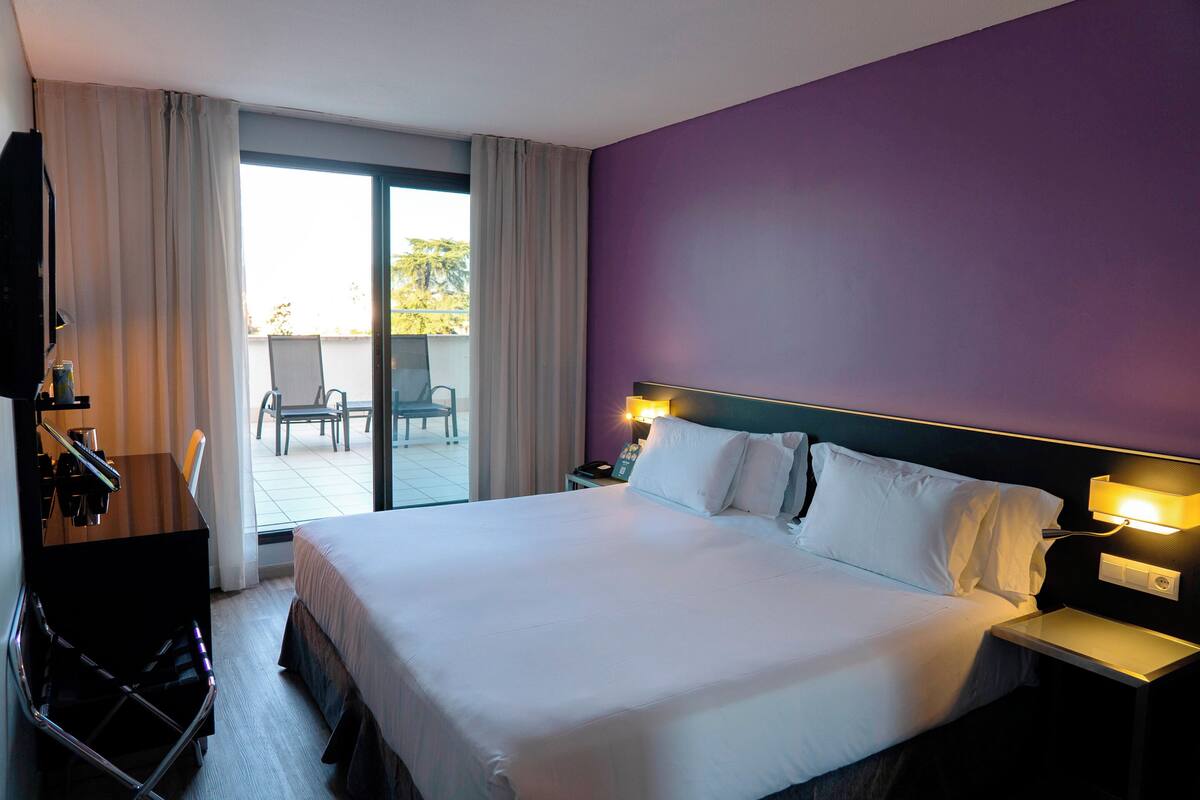 A hotel room with a large bed, a desk with a chair, a TV, and a balcony with a chair and table.