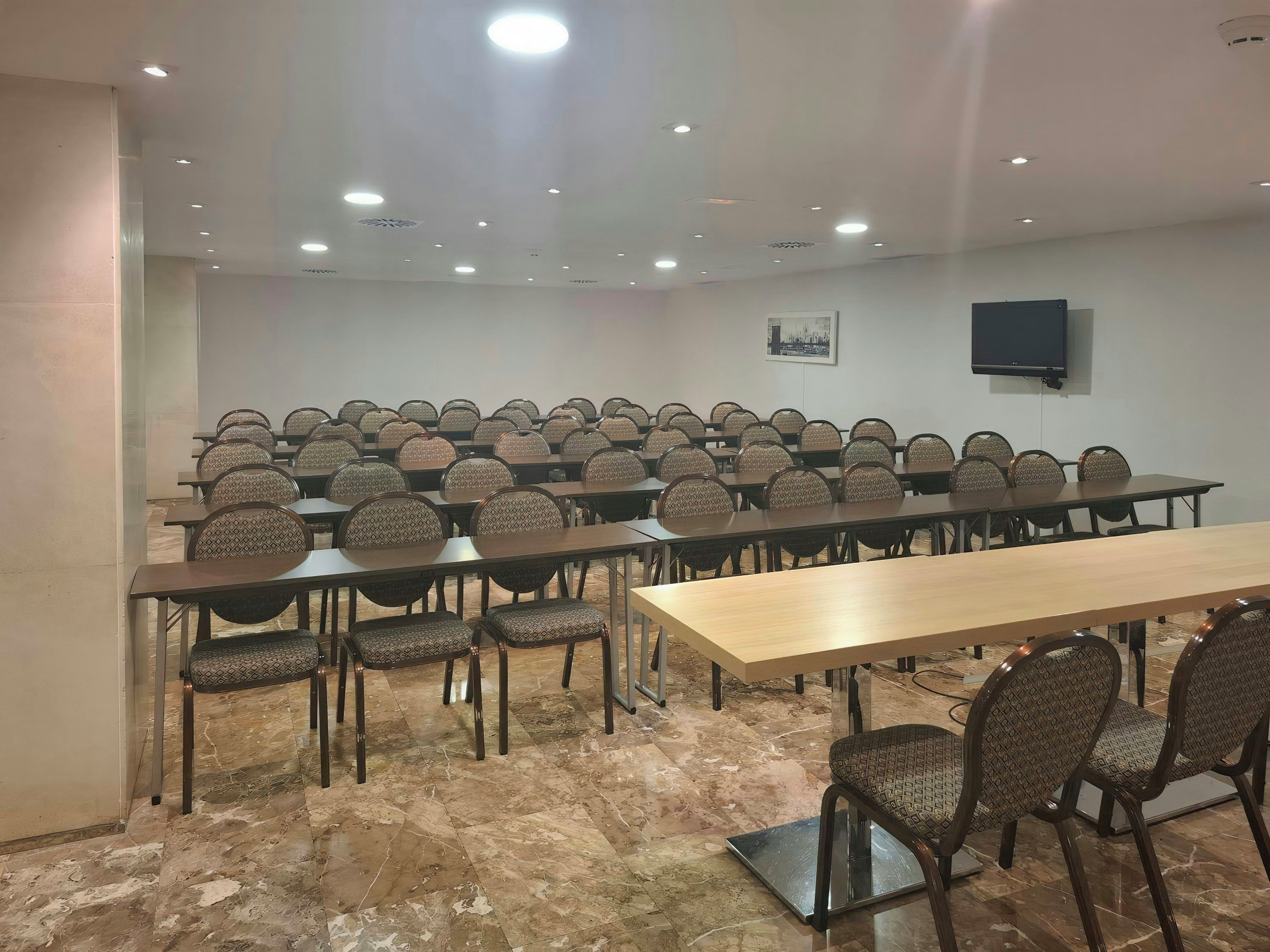 A conference room with rows of tables and chairs, a flat-screen TV mounted on the wall, and a marble floor.