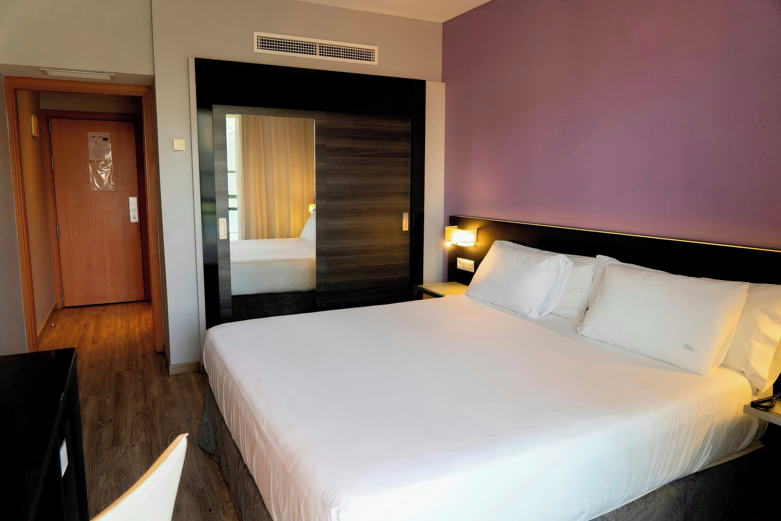 Habitación de hotel con una cama grande, una mesita de noche con lámpara, una puerta de madera y una ventana con cortinas.