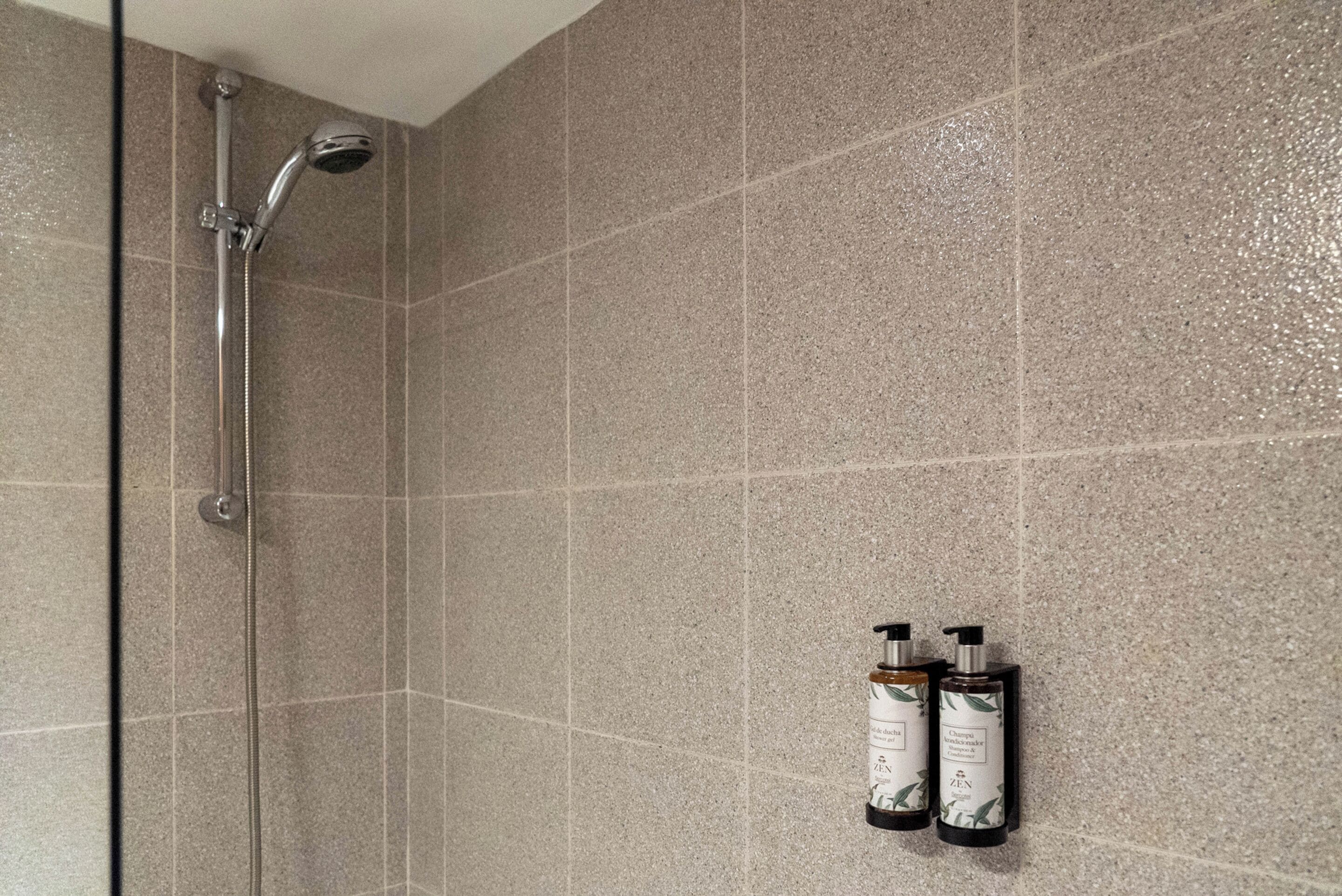 Zona de ducha con puerta de vidrio, cabezal de ducha manual y dos botellas de champú y acondicionador en la pared.