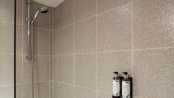 Zona de ducha con puerta de vidrio, cabezal de ducha manual y dos botellas de champú y acondicionador en la pared.