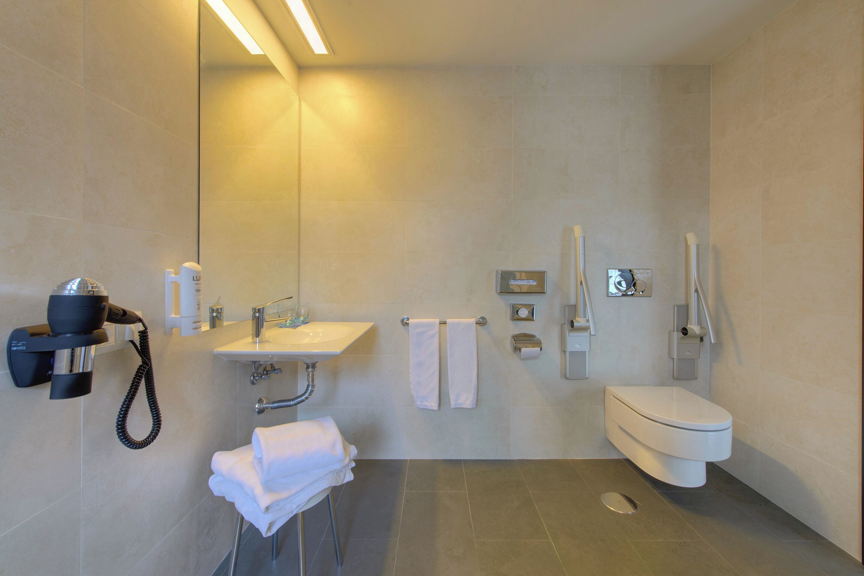 A modern bathroom with a sink, toilet, and shower. There are towels and a hairdryer on the wall.