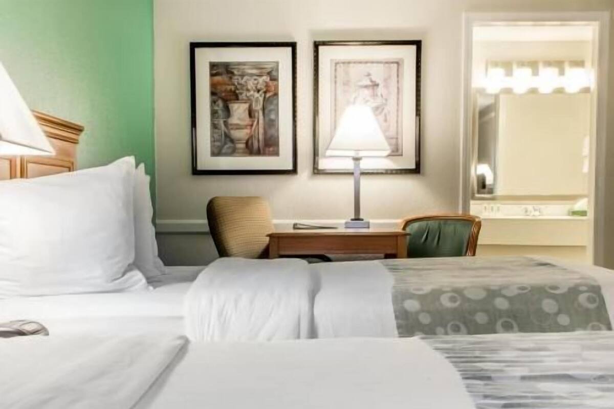 A hotel room with a bed, a desk, two framed artworks on the wall, and a window with a view.