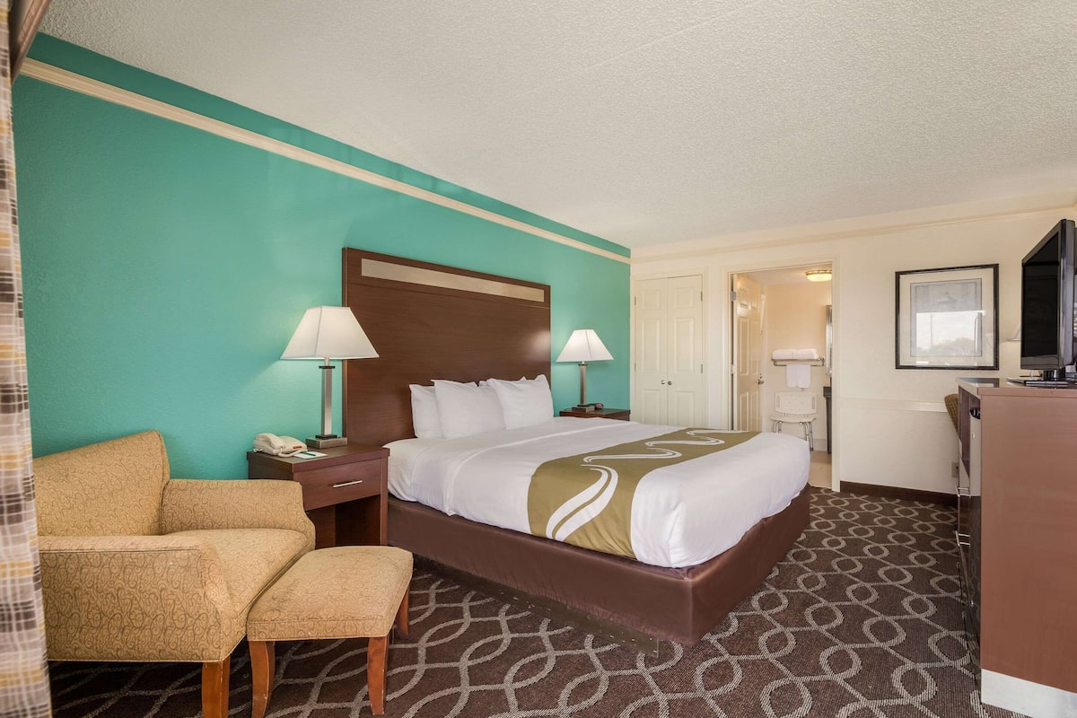 A hotel room with a bed, two bedside lamps, a desk, a chair, a TV, and a door leading to another room.