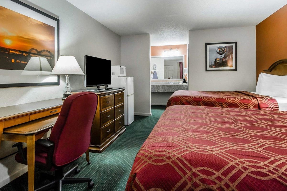 Quarto de hotel com escrivaninha, televisão, micro-ondas, cadeira vermelha, duas camas, espelho grande e obras de arte nas paredes.