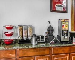 Una encimera de cocina con un fabricante de gofres, una cafetera y una plancha para gofres. También hay dos tazones rojos y una caja de cereales.
