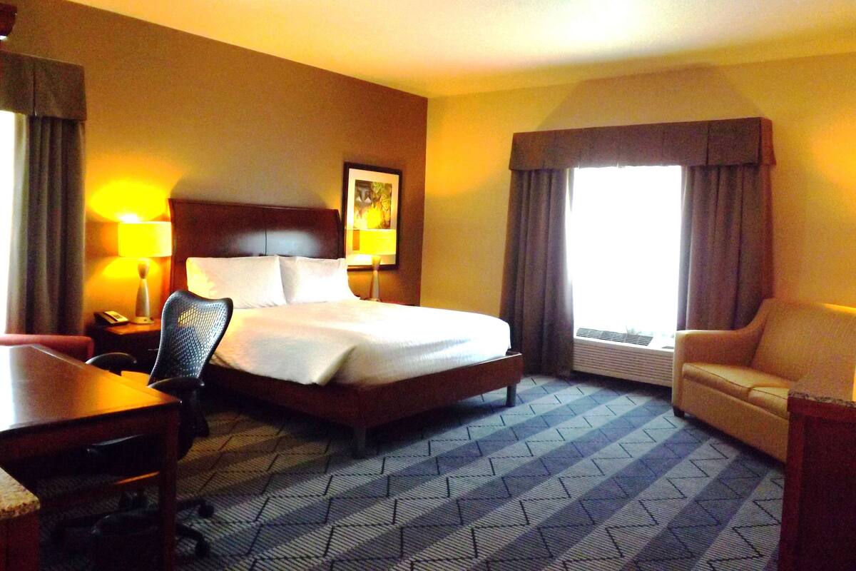 A hotel room with a bed, a desk, a chair, a lamp, and a window with curtains.
