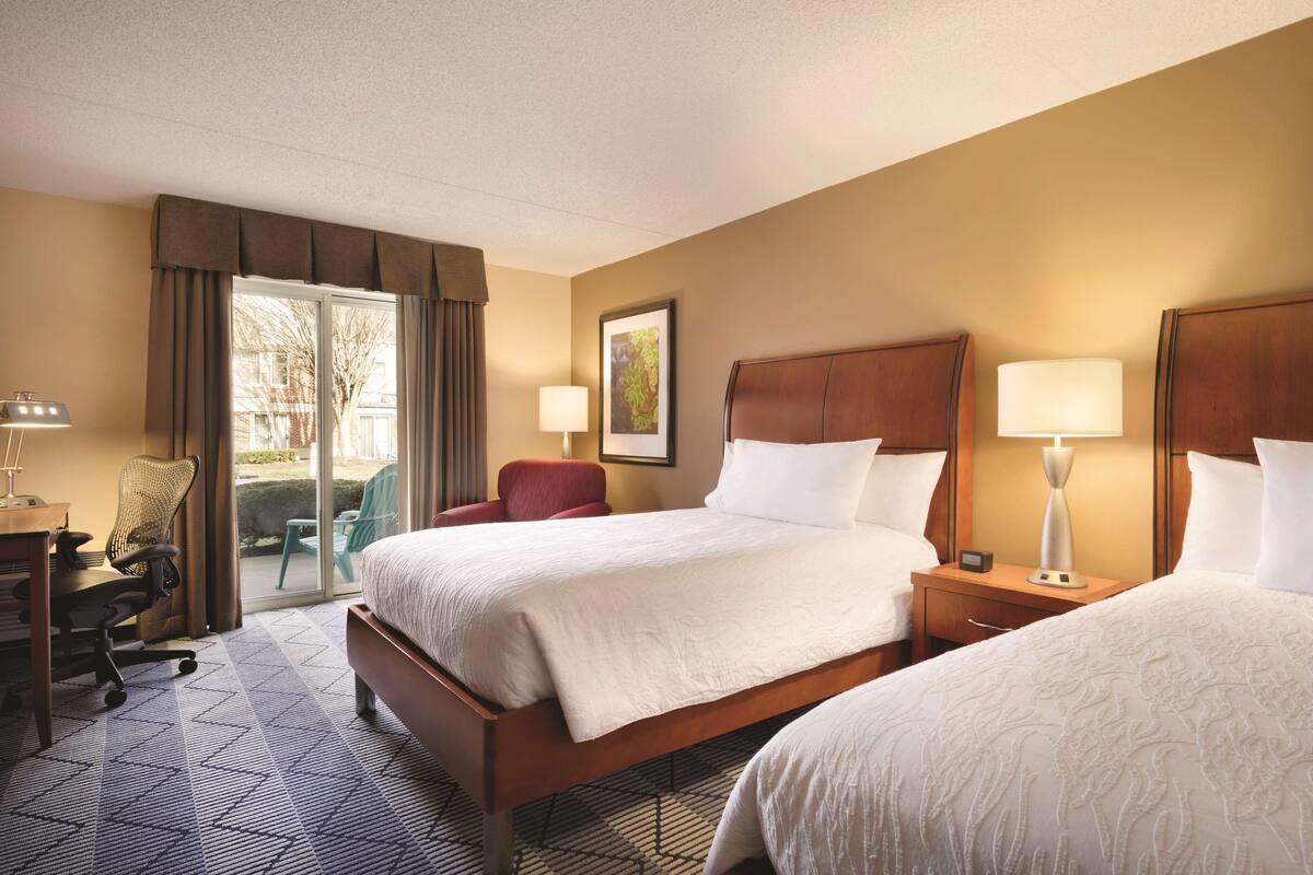 A hotel room with two beds, a desk with a chair, a lamp, and a window with curtains.