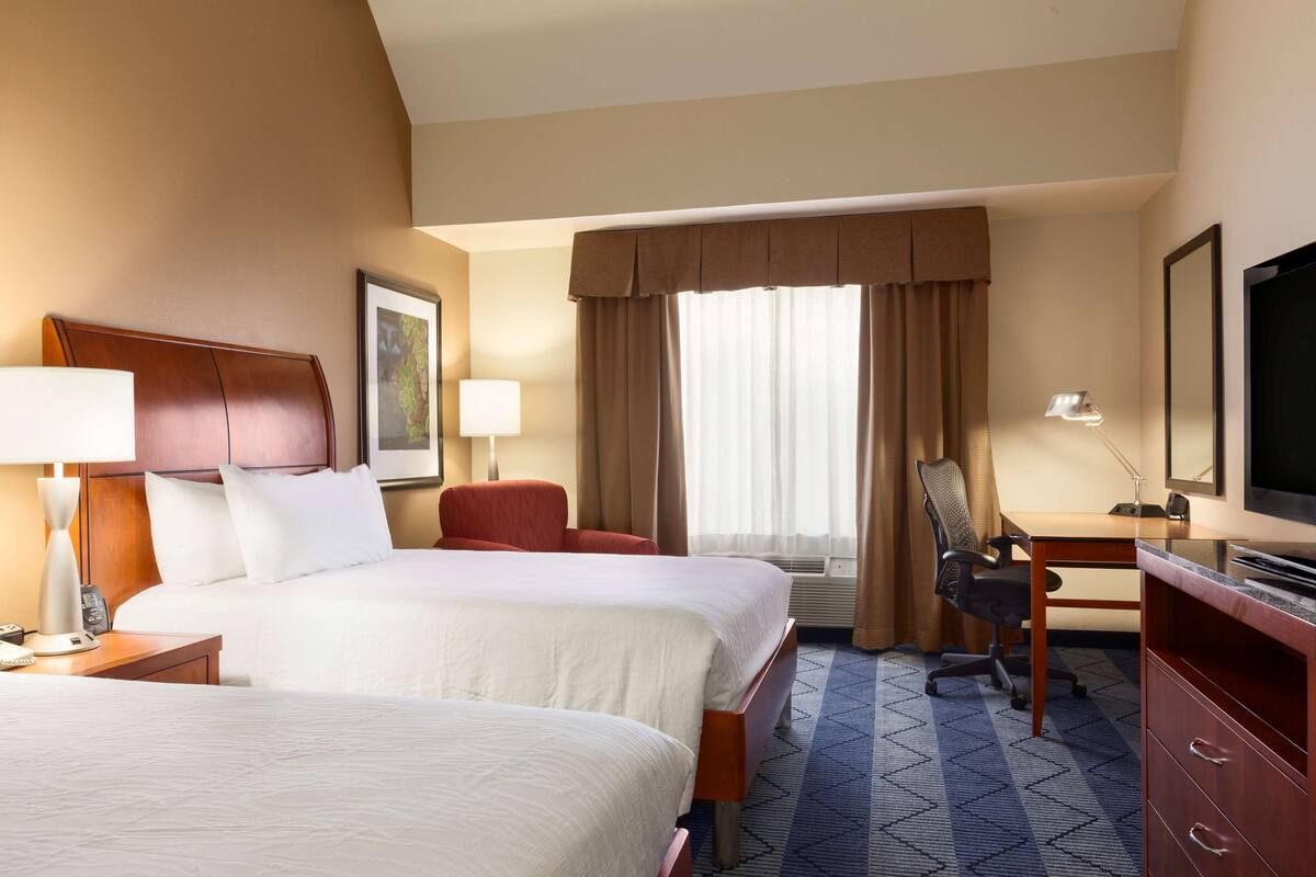 A hotel room with two beds, a desk, a chair, a television, and a window with curtains.