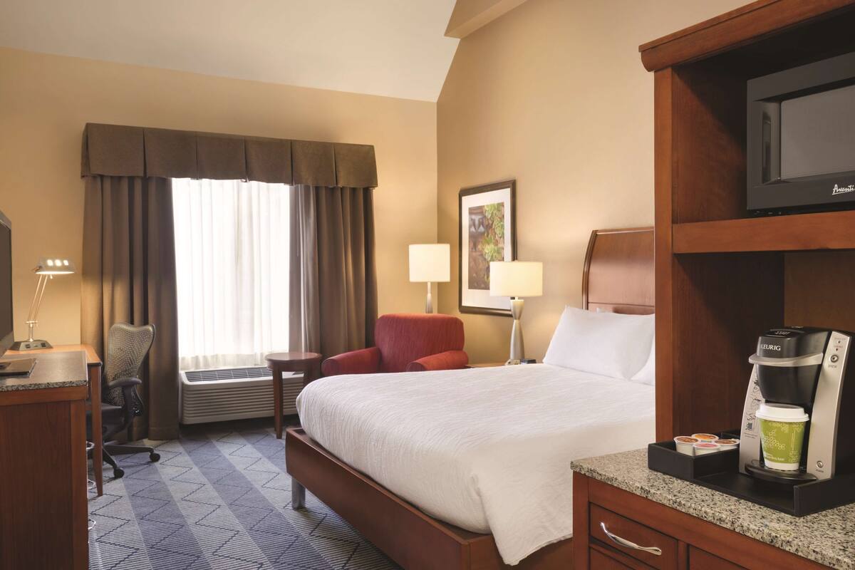 A hotel room with a bed, a desk, a chair, a coffee maker, and a window with curtains.