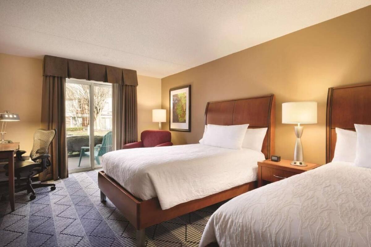 A hotel room with two beds, a desk, a chair, and a view through a glass door.