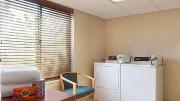 A laundry area with a washing machine, a chair, a table with laundry detergent, and a window with blinds.