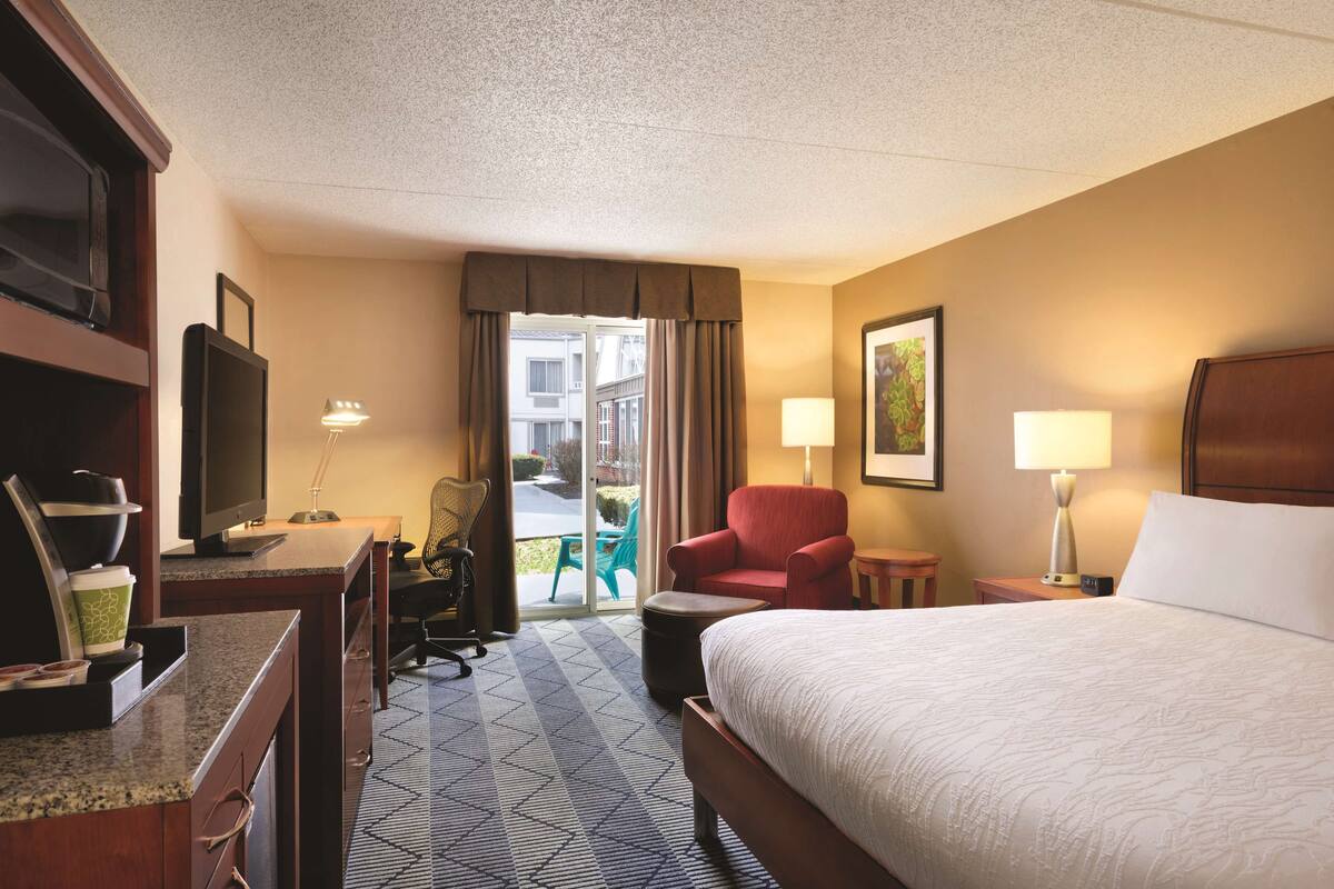 A hotel room with a bed, a desk with a television, a chair, a lamp, and a view of the outside.